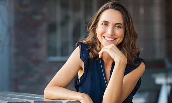 a woman with long brown hair is sitting on a couch smiling brightly she is wearing a white sleeveless top and the background is softly blurred focusing attention on her joyful expression the lighting is warm and highlights her cheerful demeanor subtly enhancing the results of recent cosmetic surgery