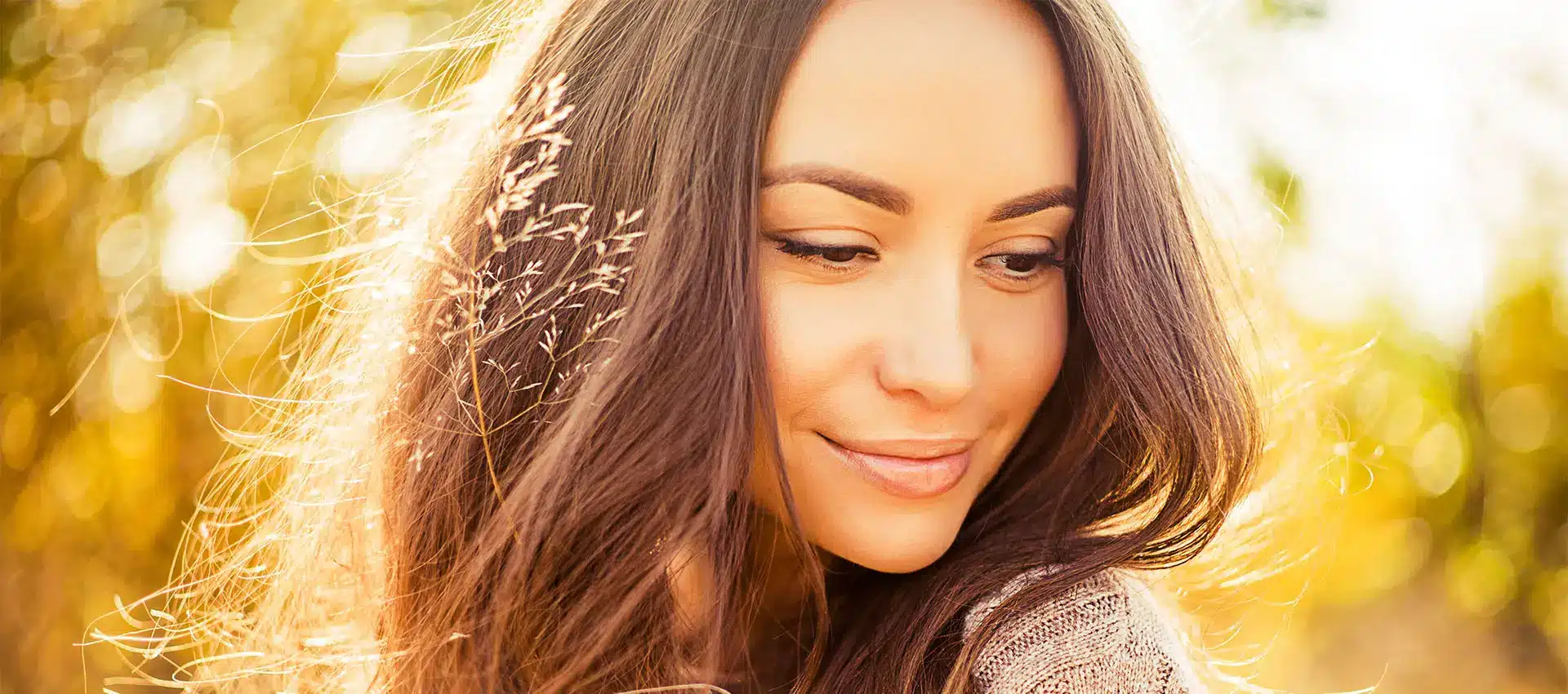 a woman with long brown hair having recently had an eye lift smiles gently while looking down she is outdoors surrounded by a warm and sunny background with a golden glow that causes her hair to reflect the light