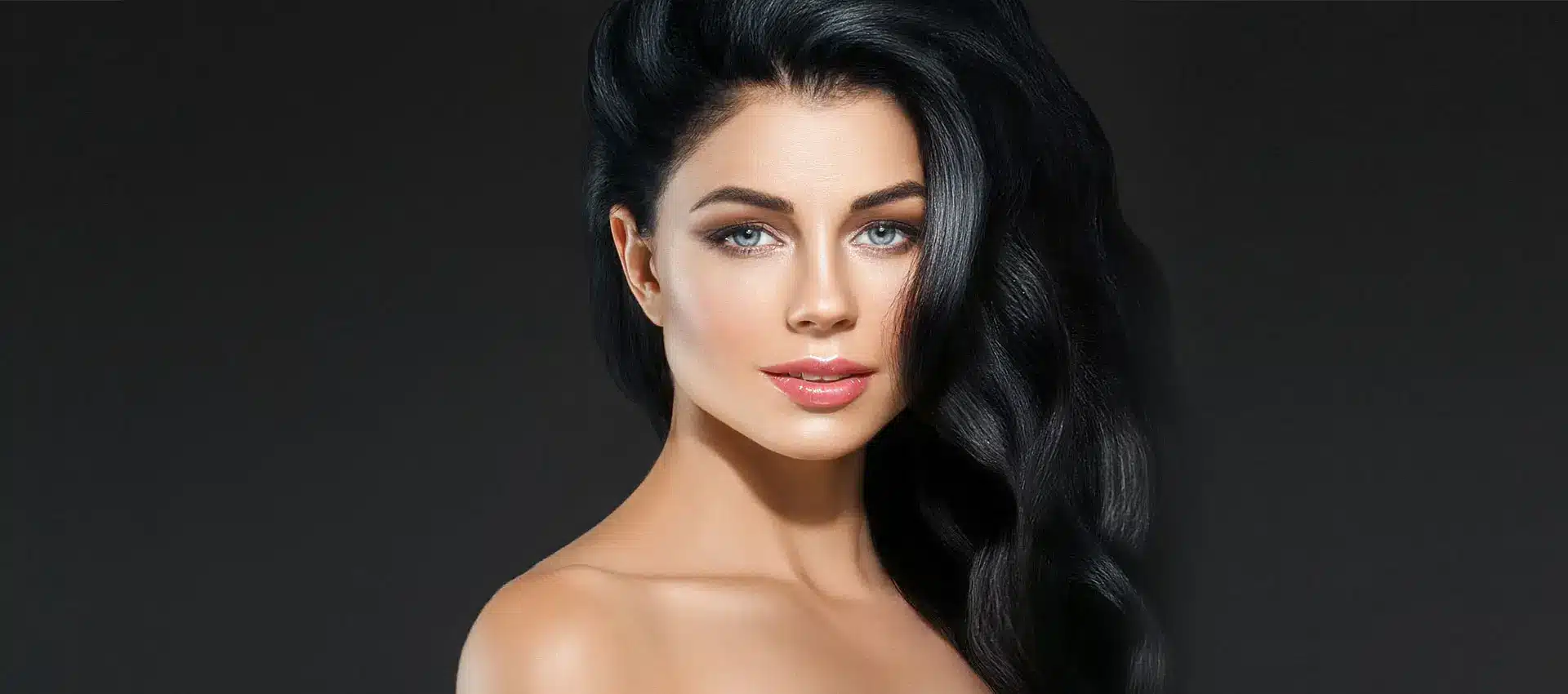 portrait of a woman with long wavy black hair draped over one shoulder against a dark background she has blue eyes light makeup and a neutral expression gazing directly at the camera her shoulders are bare emphasizing her elegant and composed appearance subtly enhanced by a brow lift