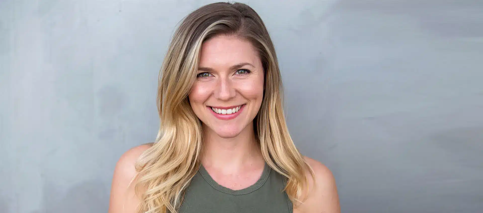 smiling individual with long blonde hair wearing a sleeveless green top stands against a gray background showcasing the impressive results of facetite skin tightening and facial contouring