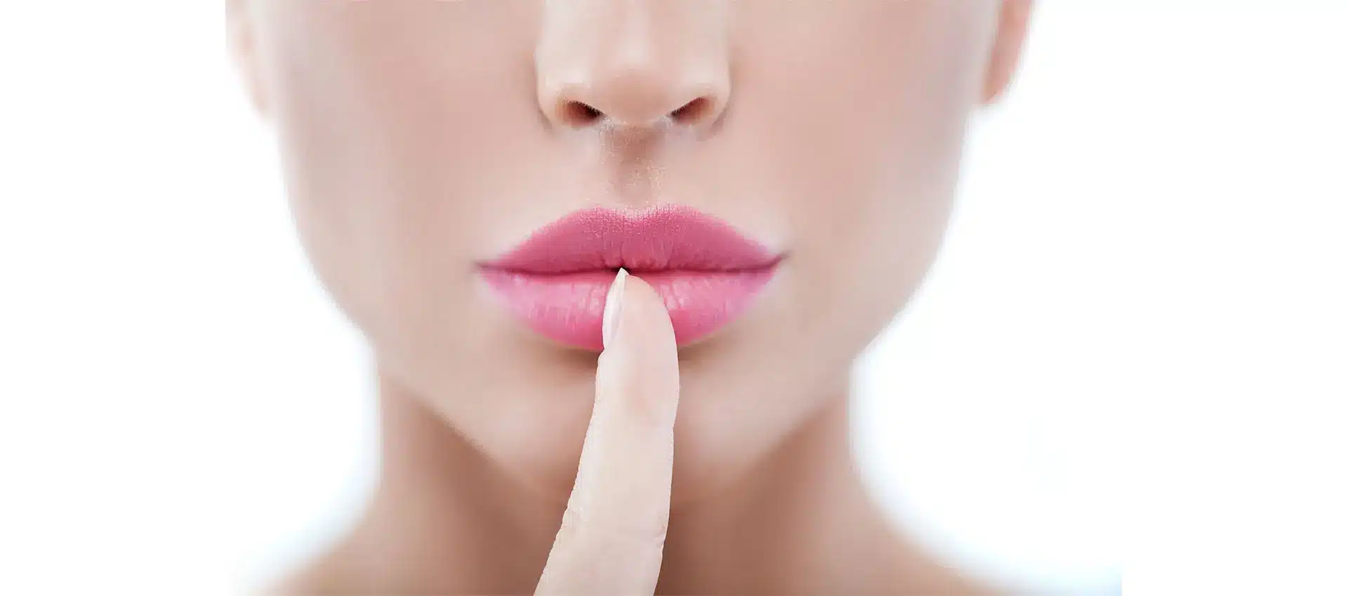 close up of a person with pink lips possibly enhanced by a recent lip lift holding their index finger vertically in front of their mouth suggesting they are asking for silence or secrecy the background is white and slightly blurred