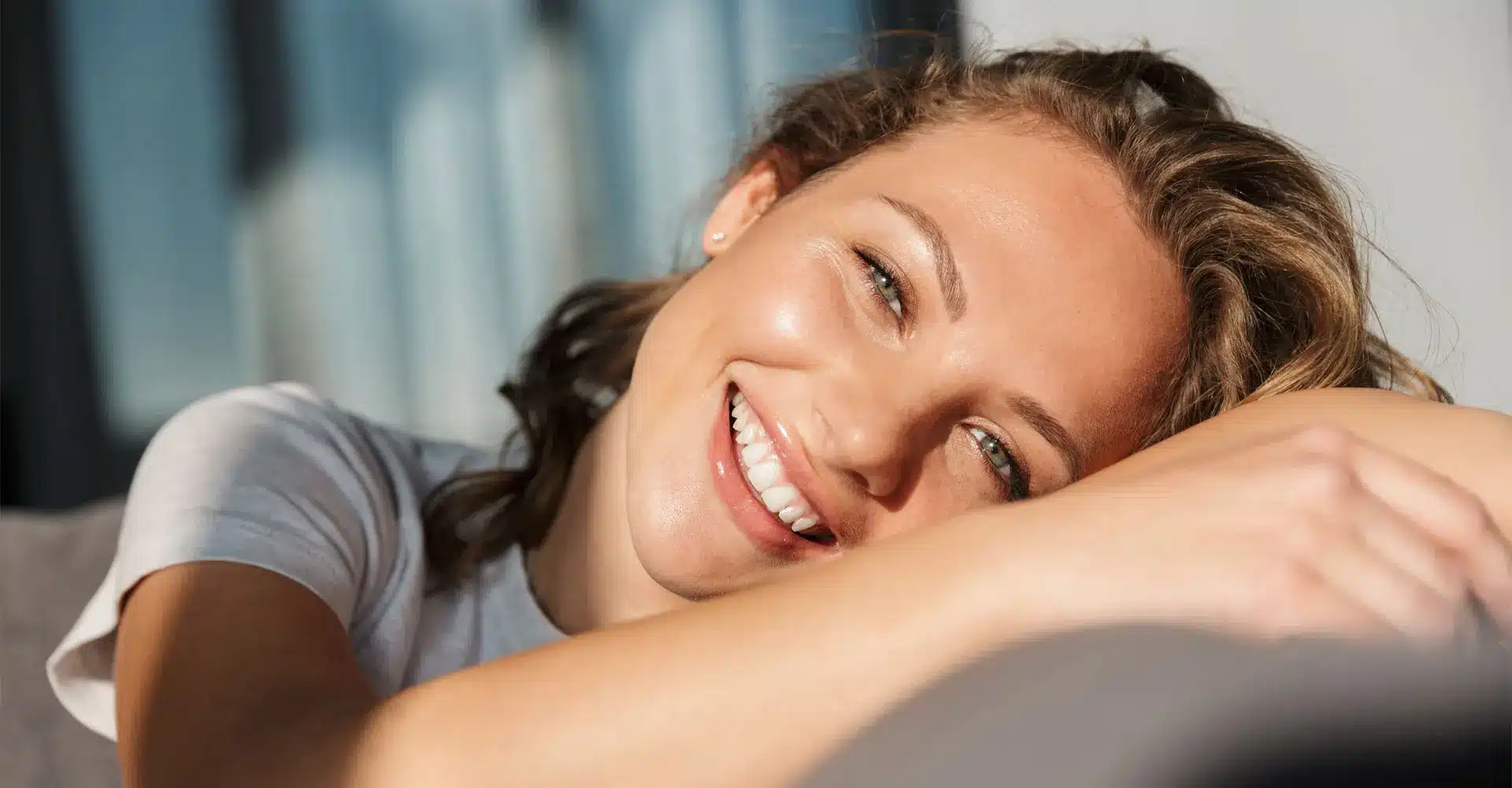 a smiling person with light brown hair is leaning on their arm bathed in natural sunlight they are wearing a white shirt and appear relaxed and content the background possibly suggesting a family treatment session is slightly blurred and features soft neutral tones