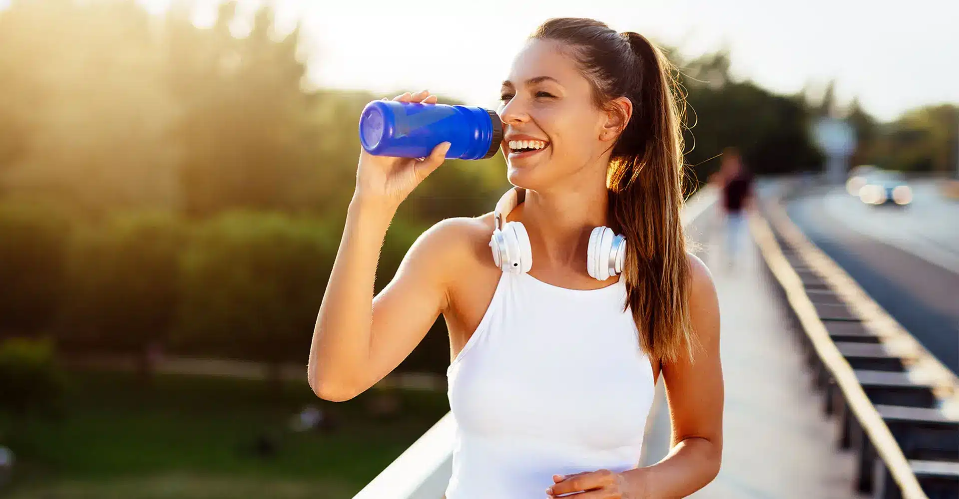 a woman in athletic wear with headphones around her neck is smiling and drinking from a blue water bottle she is outdoors on a sunny day standing on a pedestrian bridge or path with greenery in the background looking refreshed after her morning workout and successful kybella treatment