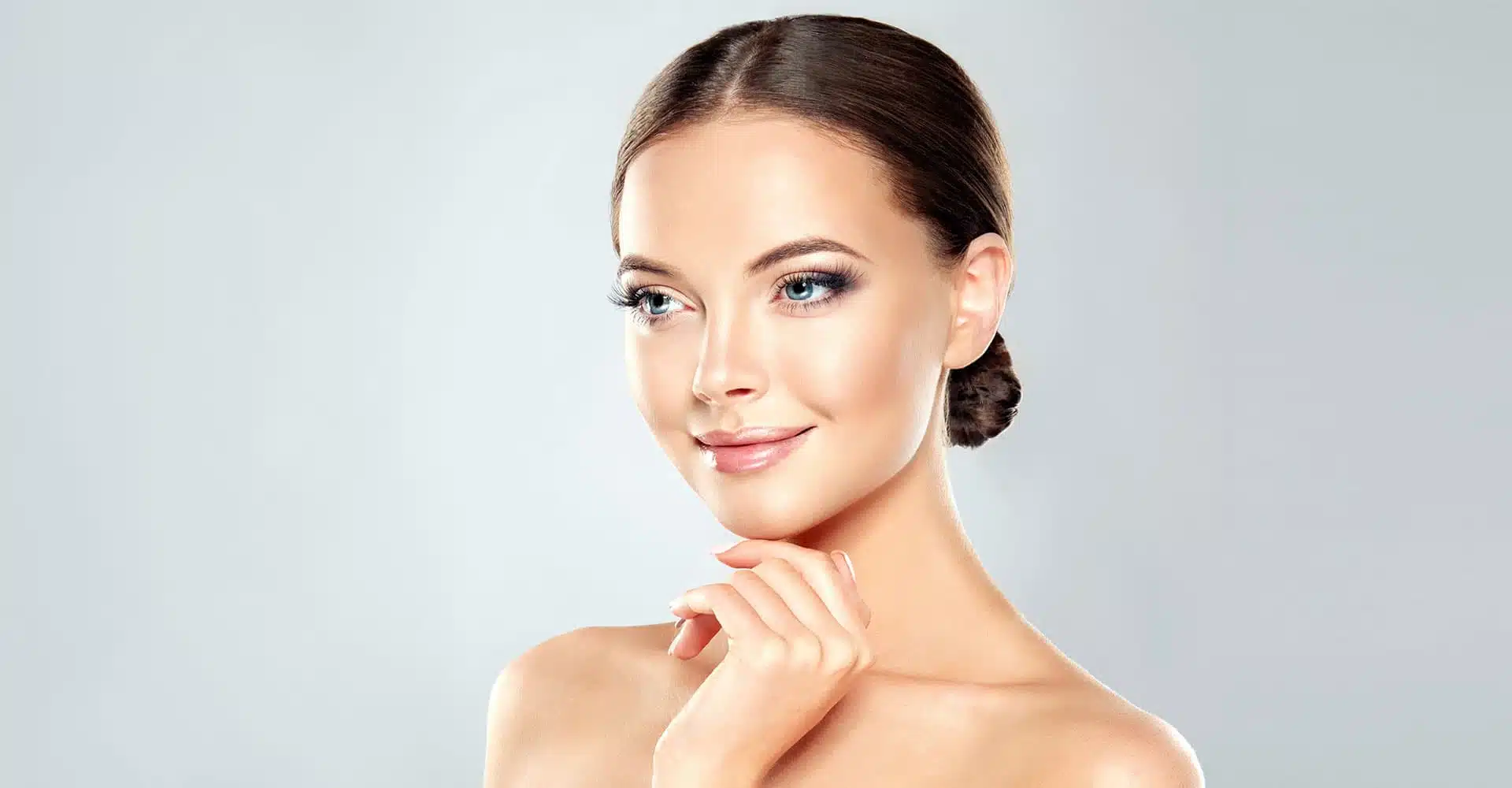 a woman with a smooth complexion and sleek dark hair pulled back into a low bun is looking to the side with a slight smile she is touching her chin lightly with one hand showcasing minimalistic makeup that highlights her features possibly benefitting from non surgical rhinoplasty set against a light gray background