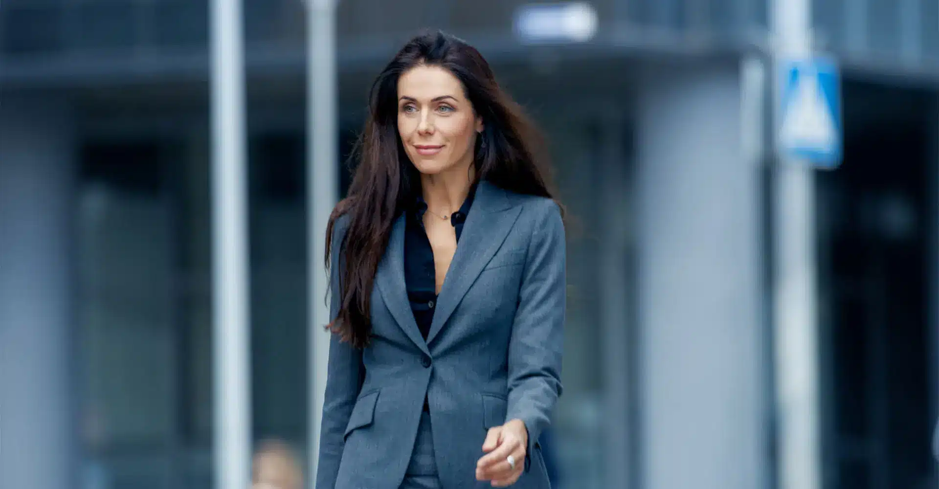 a woman with long dark hair wearing a gray suit over a black top walks confidently outdoors the background is blurred featuring a modern building and some street elements her radiant skin hints at the subtle benefits of cosmetic treatment