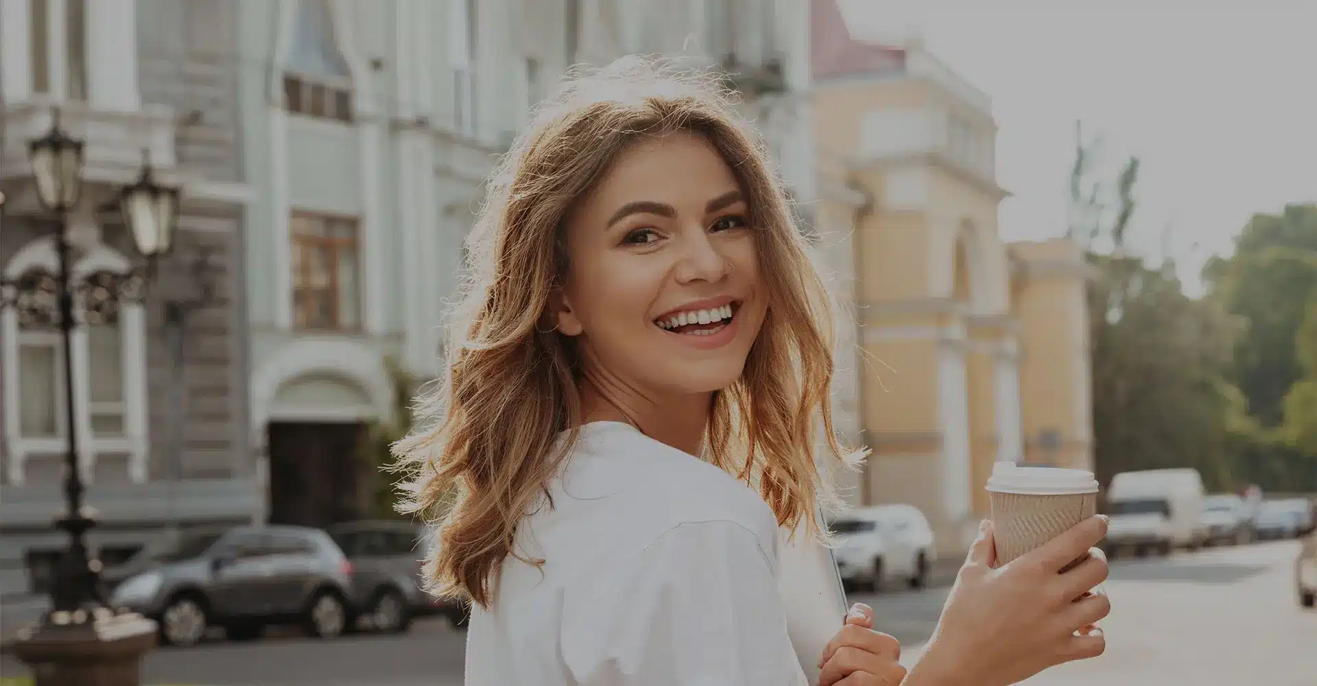 a young woman with shoulder length light brown hair is smiling and looking back at the camera while holding a takeaway coffee cup she is outdoors on a sunny day standing in front of elegant buildings and parked cars subtly showcasing the glow of recent facial rejuvenation she wears a casual white shirt
