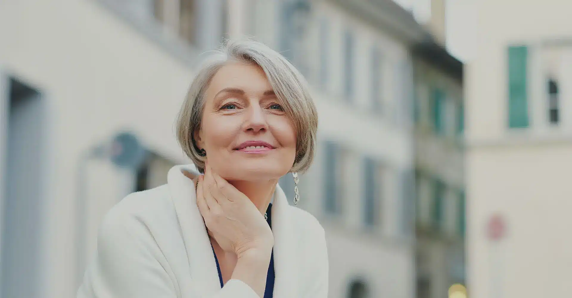 a woman with short gray hair smiles softly while gently touching her neck perhaps considering neuromodulators she wears a white jacket and a pair of dangling earrings the blurred background features buildings in a casual urban setting