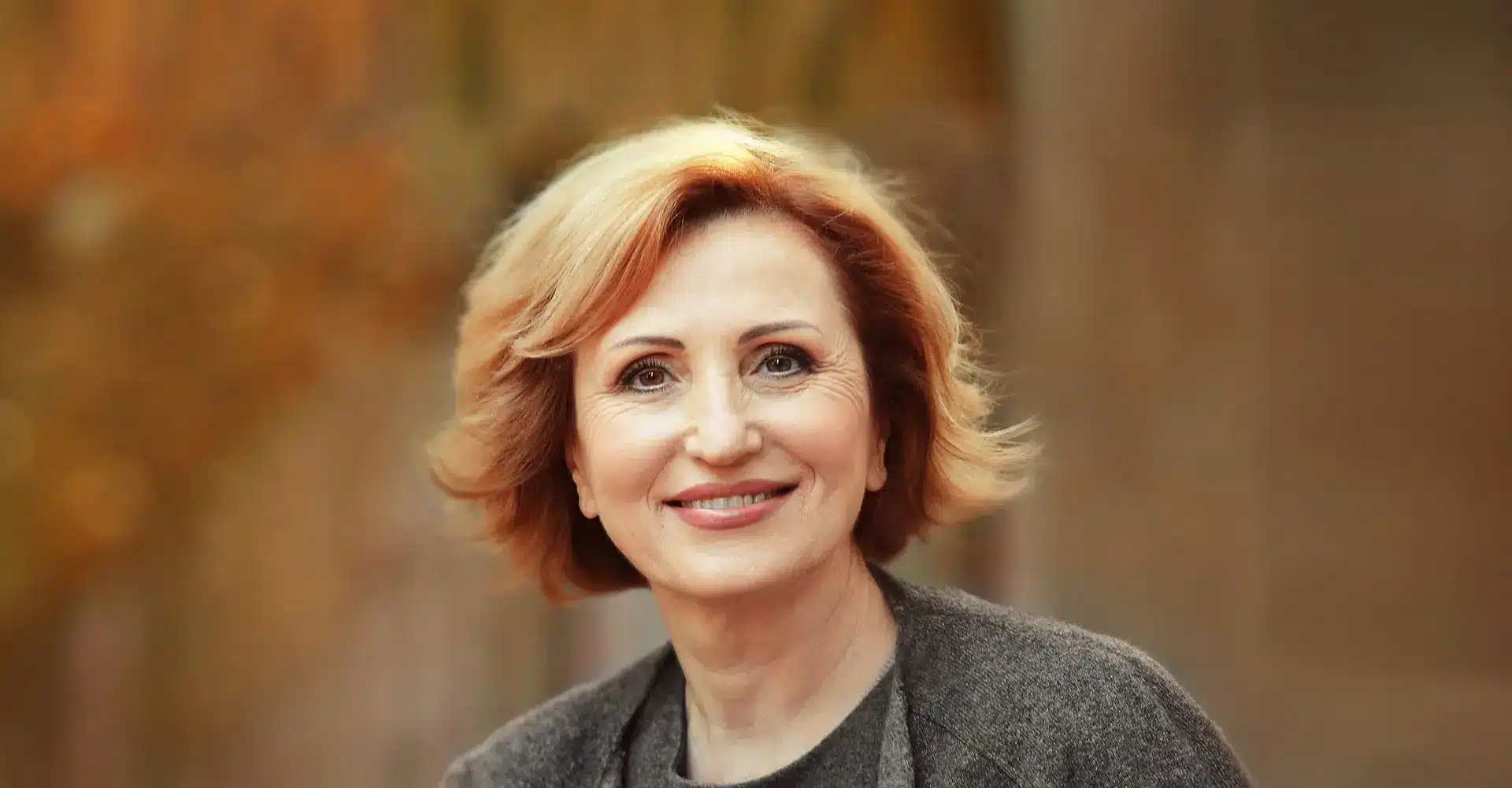 a woman with short wavy blonde hair smiles warmly she is wearing a gray sweater and is set against a blurred background of autumn colors giving the image a cozy and serene feel her bright eyes appear even more rejuvenated after a tear trough augmentation