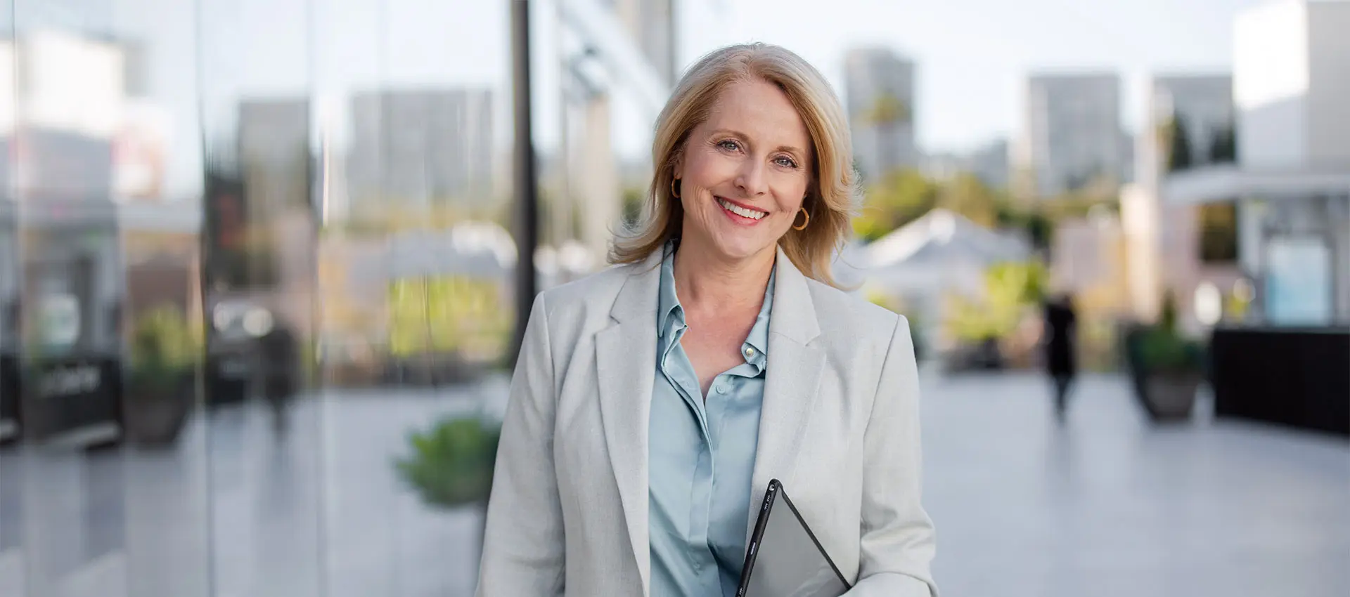 a smiling woman with blonde hair wearing a light grey blazer and carrying a tablet stands outdoors in a modern urban setting with blurred buildings in the background her youthful radiance perhaps enhanced by neuromodulators