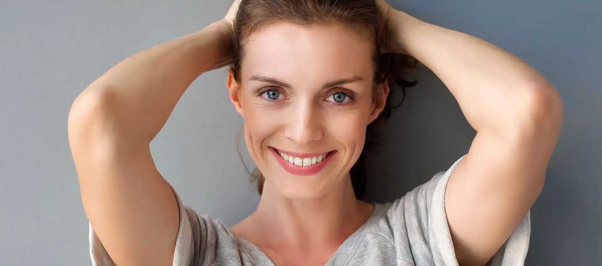 a woman with light skin and brown hair smiles at the camera she wears a light grey top and has her arms raised hands behind her head against a neutral background her radiant complexion hints at a recent juvederm treatment which enhances her natural beauty effortlessly