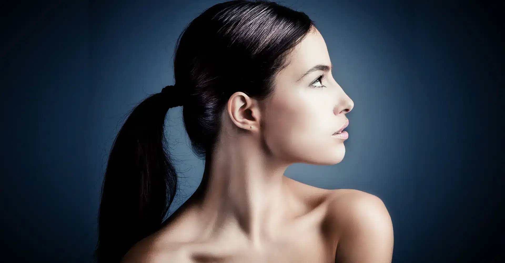 a young woman with dark hair tied in a ponytail is shown in profile against a dark gradient background her bare shoulders are visible and she gazes intently to her left the image emphasizes her smooth complexion elegant neck and the refined contour achieved through kybella cosmetic treatment