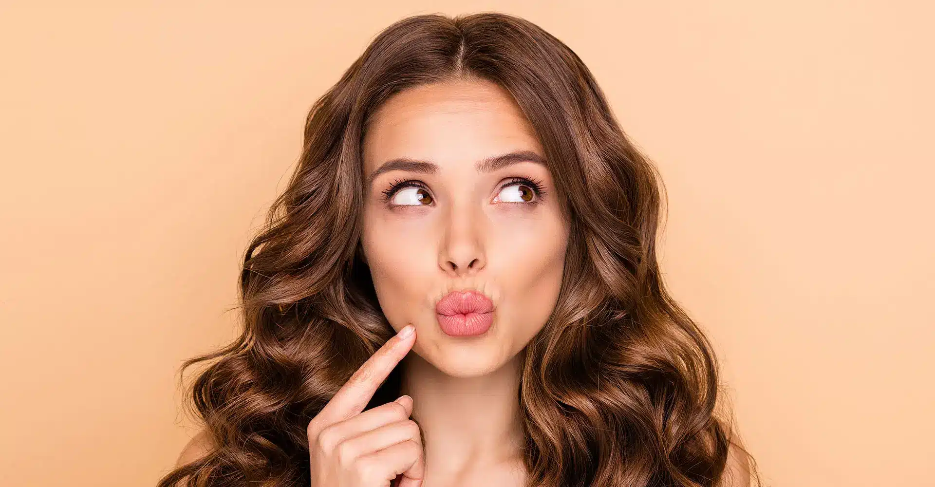 a woman with long wavy brown hair is standing against a peach colored background she has a thoughtful expression looking up and to the side with her lips puckered perhaps showcasing recent lip enhancement as her finger rests on her cheek