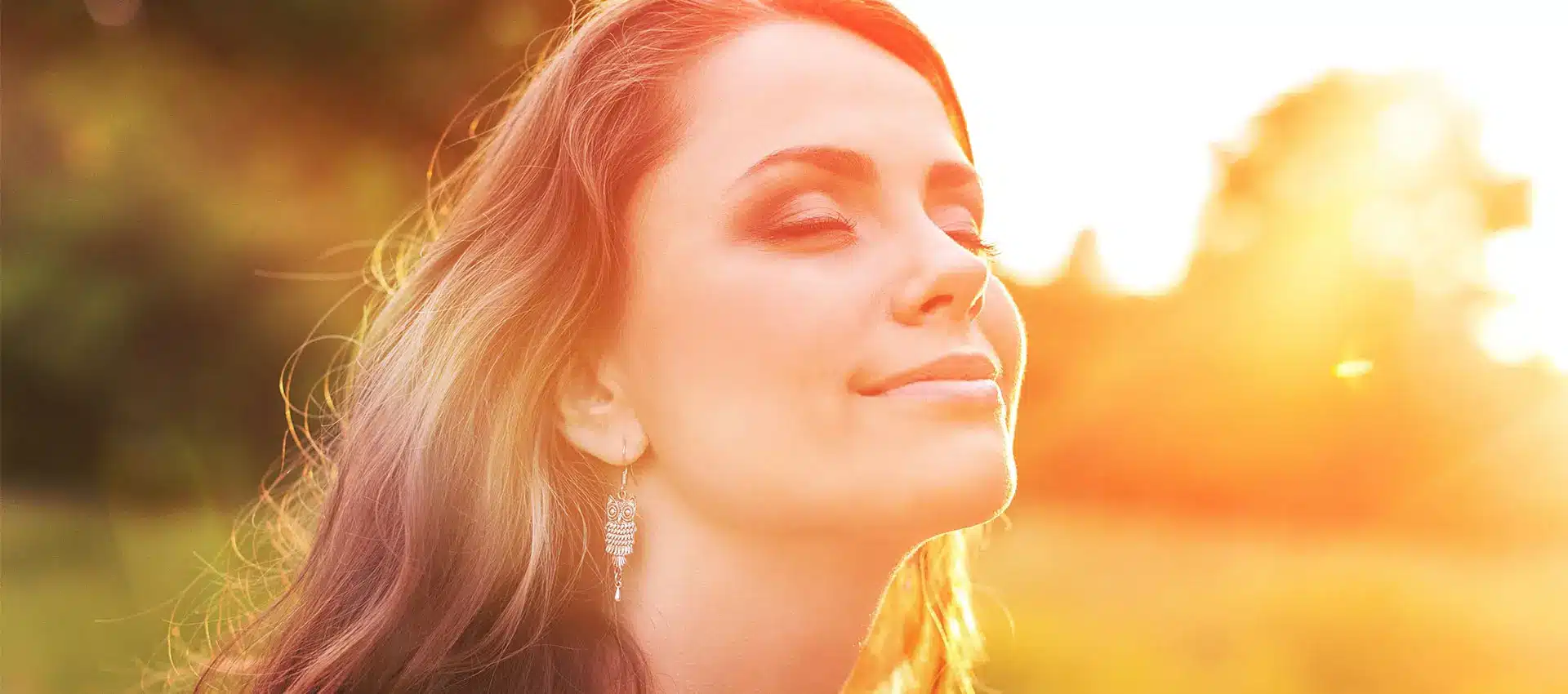 a woman with long brown hair and silver earrings is outdoors with her eyes closed basking in the sunlight the background is blurry with warm sunlight illuminating the scene creating a serene and peaceful ambiance her skin glows as if enhanced by restylane adding to her natural beauty