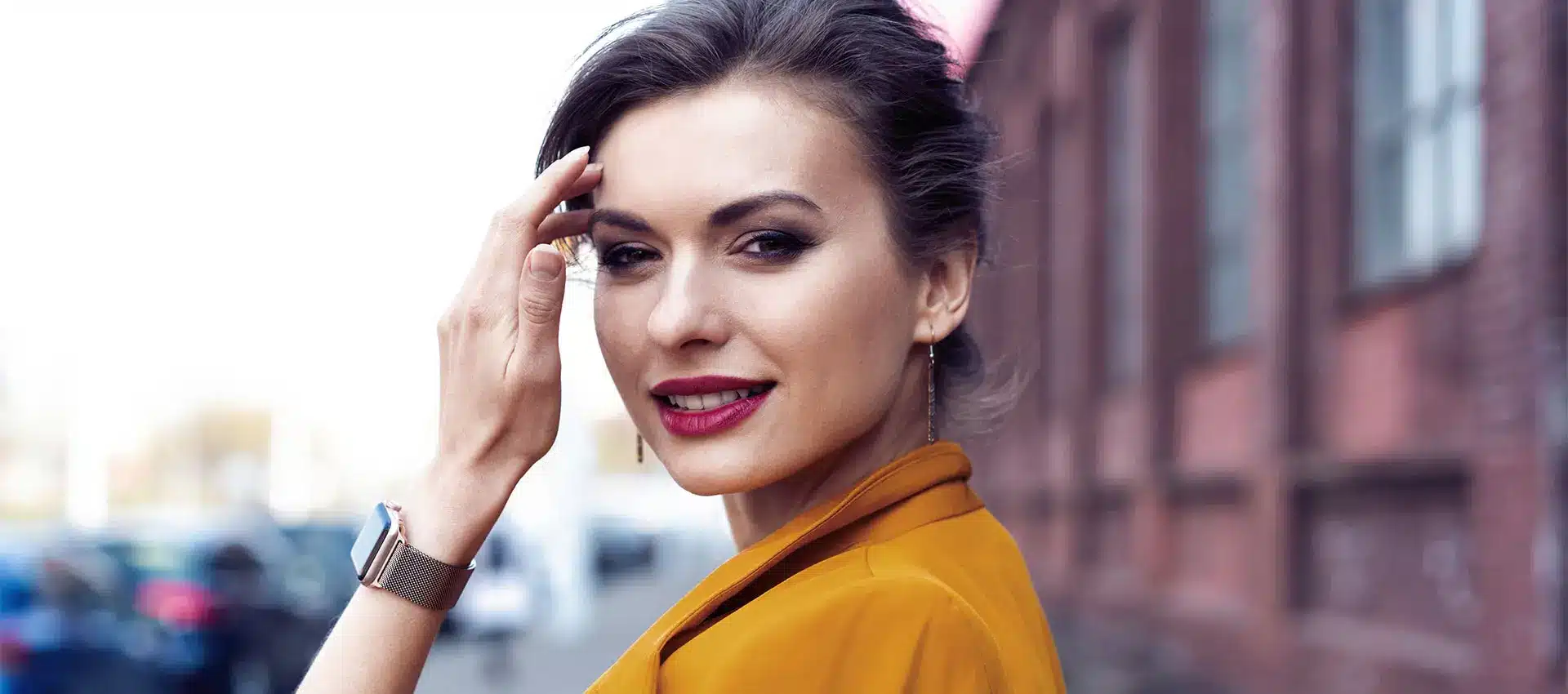 a woman with dark hair pulled back wearing a mustard yellow jacket stands outdoors with a confident expression delicate earrings and a smartwatch adorn her wrist the background slightly out of focus reveals a brick building and some vehiclespossibly after an enjoyable family treatment outing
