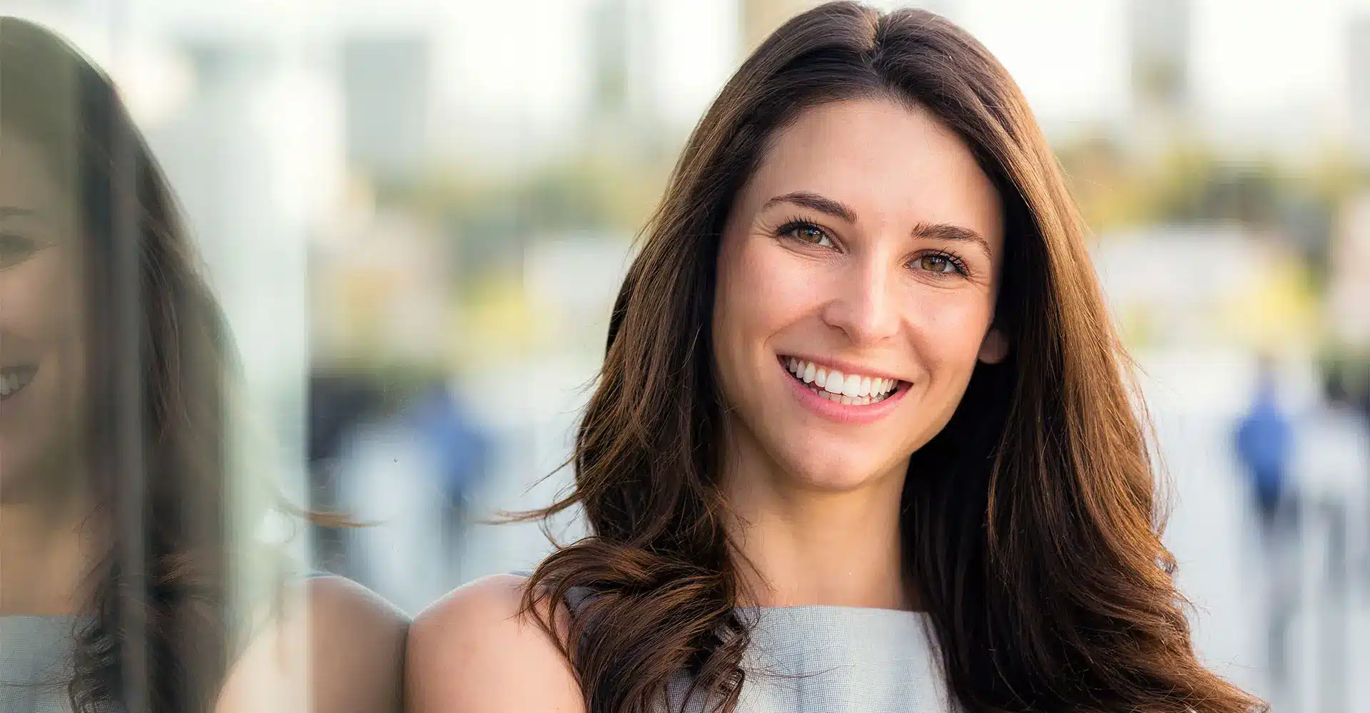 a woman with long brown hair smiles warmly at the camera she is standing outdoors with a cityscape blurred in the background and her reflection is visible on a glass surface beside her after a non surgical nose job the subtle change enhances her natural beauty the overall atmosphere is bright and cheerful
