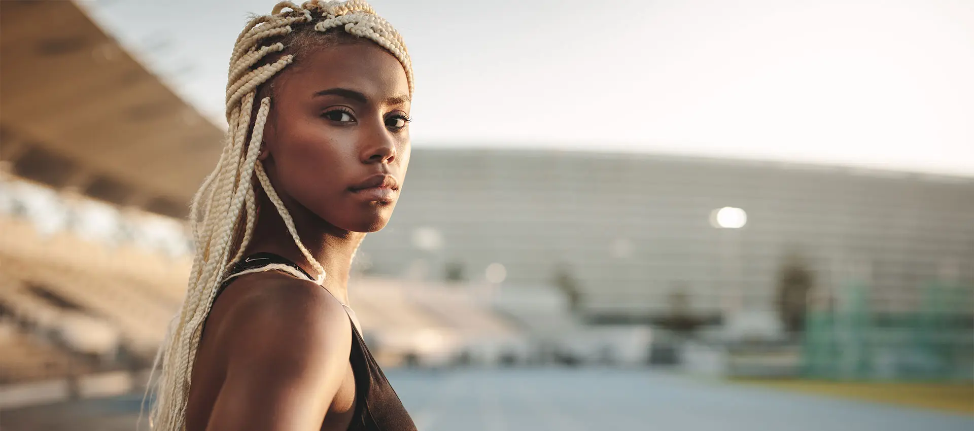 a woman with braided blonde hair stands at an outdoor stadium gazing into the distance she appears focused and determined as if preparing for a competition in the background blurred bleachers and the stadium structure frame her figure the lighting suggests it is close to sunset or sunrise casting a restylane like glow on her complexion