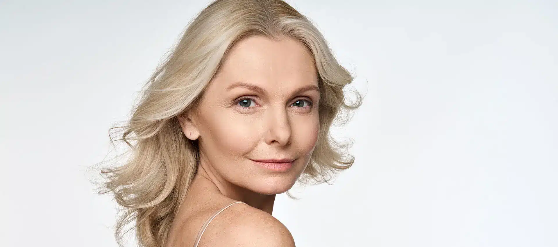 a woman with long blonde hair and light skin is looking over her shoulder towards the camera wearing a sleeveless top she has a subtle smile that radiates confidence the plain light colored background emphasizes her natural beauty reminiscent of graceful simplicity featured in seo optimized beauty campaigns