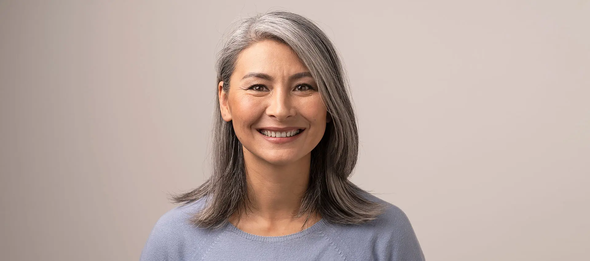 a woman with shoulder length gray hair is smiling at the camera she is wearing a light blue sweater and the background is a neutral beige color creating an inviting scene that could enhance seo for any webpage