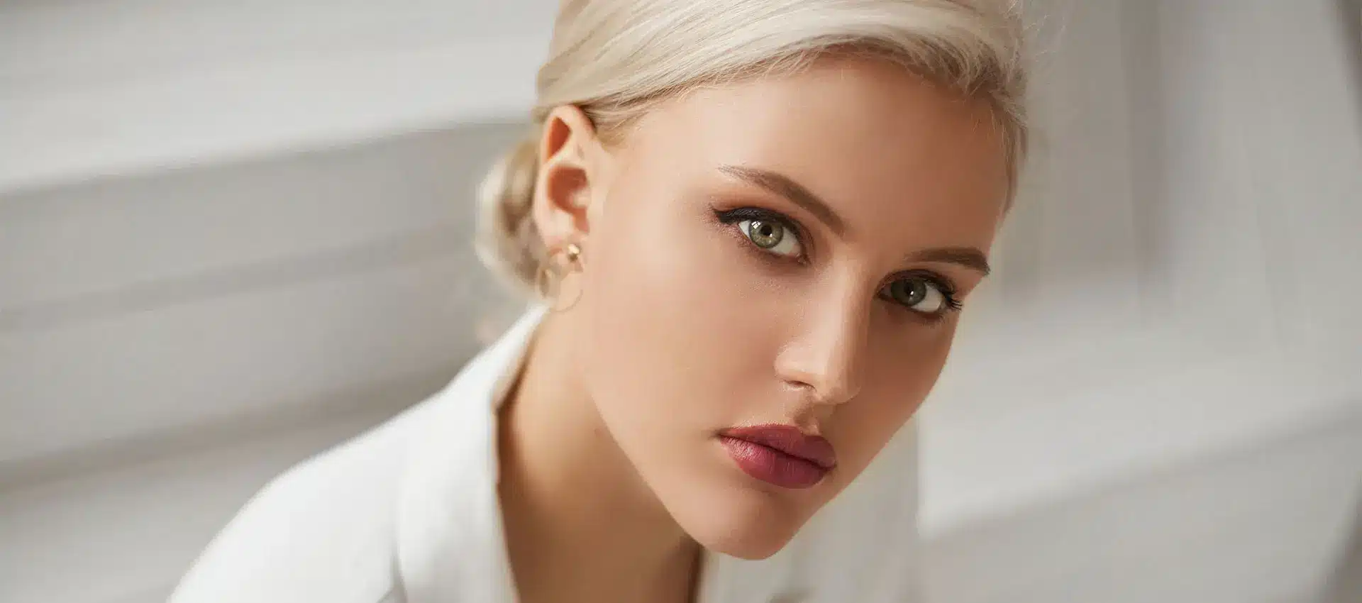 a woman with light blonde hair styled in a low bun gazes at the camera with a neutral expression she has light skin and green eyes enhanced subtly by juvederm treatments dressed in a white top and sitting against a blurred white background she emanates a serene atmosphere