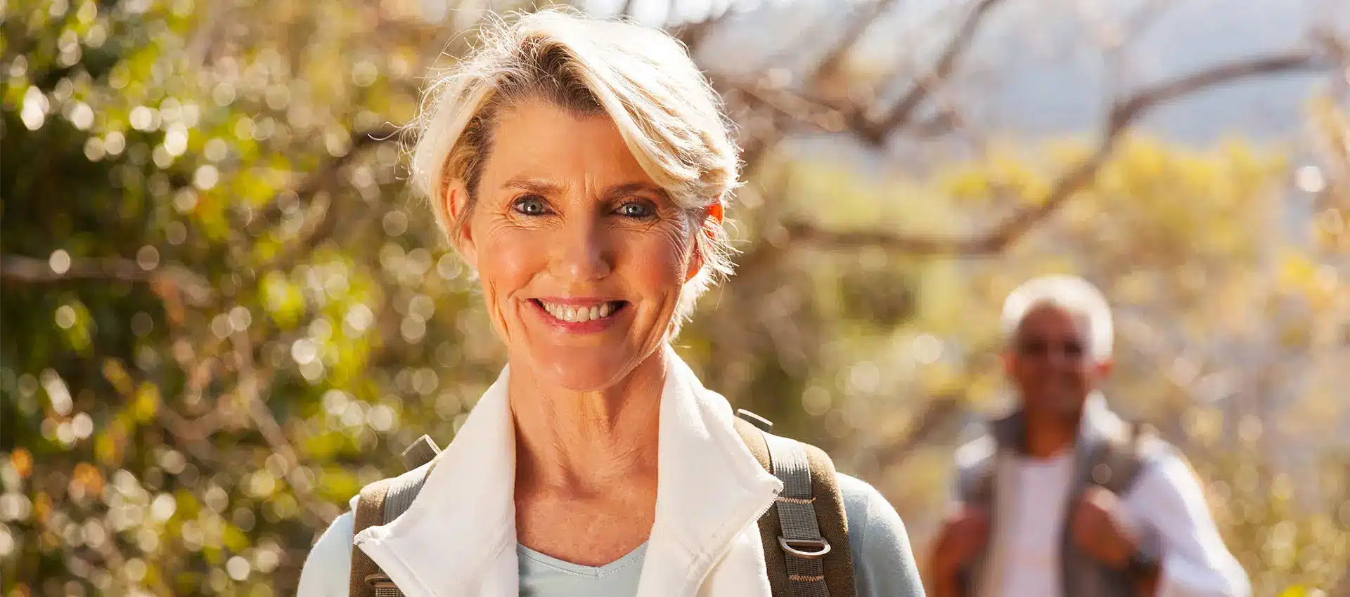 a smiling woman with short blonde hair is wearing a light blue shirt and a white vest standing outdoors with a backpack on in the blurred background an older manperhaps a family memberis also carrying a backpack set against a backdrop of trees and nature