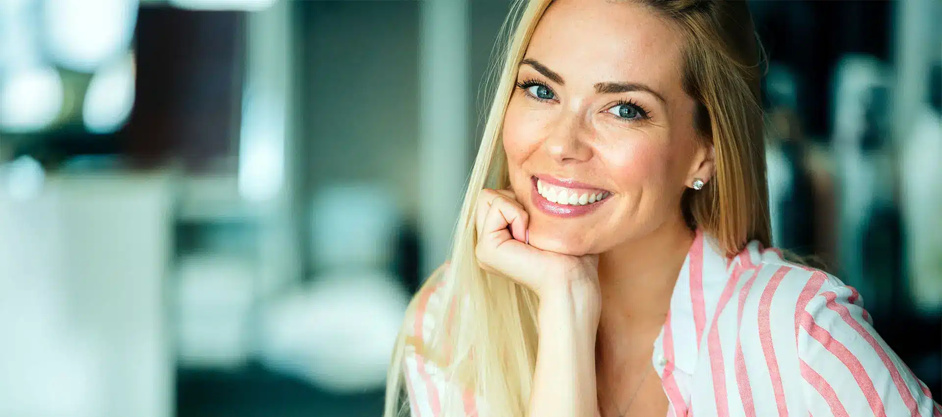 a smiling woman with long blonde hair and blue eyes rests her chin on her hand she wears a pink and white striped shirt showcasing the subtle benefits of restylane® dermal filler she sits against a blurred background creating a bright and cheerful atmosphere