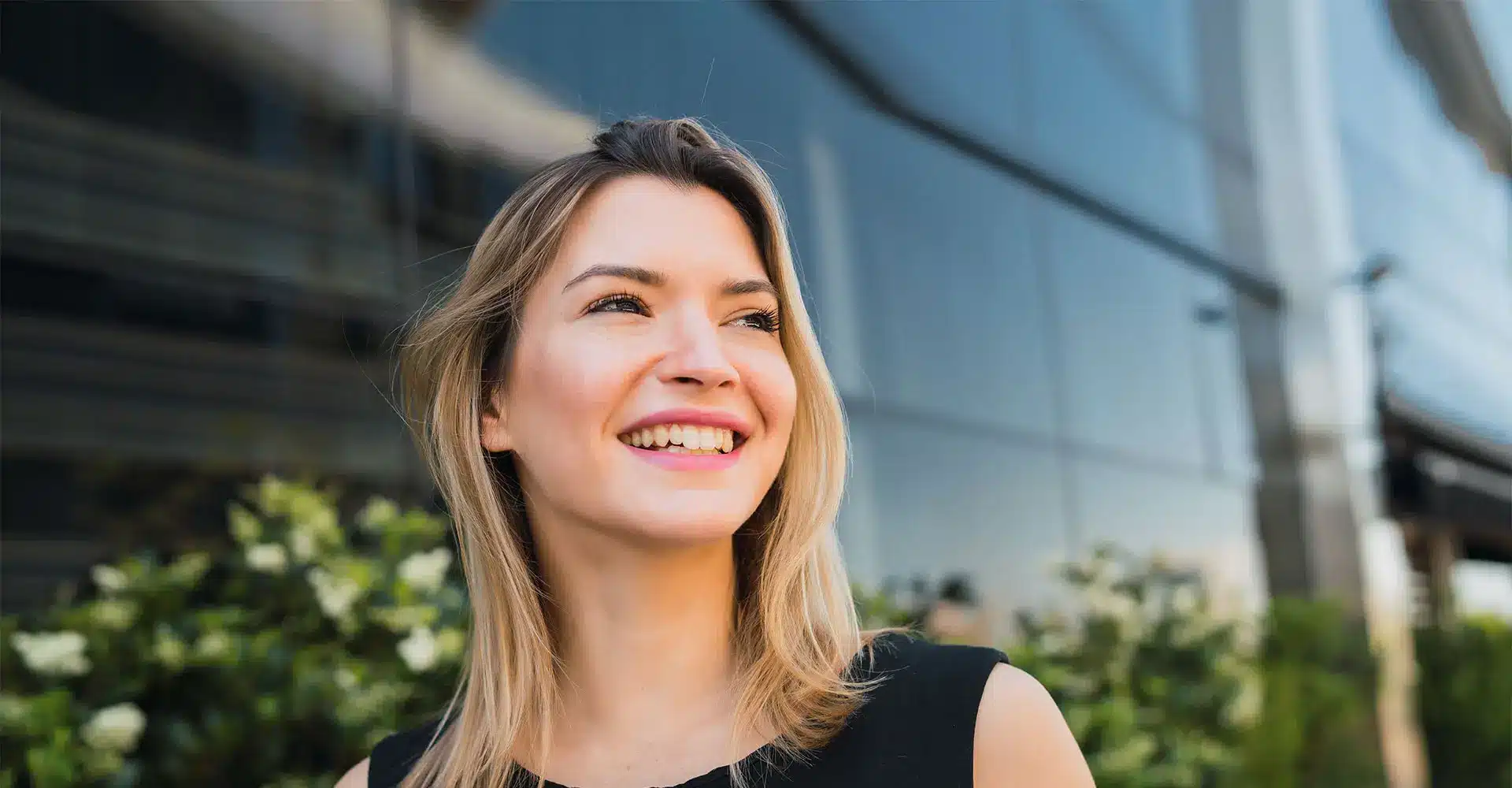 a smiling woman with shoulder length blonde hair stands in front of a glass building she is wearing a sleeveless black top and the background features some greenery and reflections on the glass her bright eyes hint at recent eyelid surgery adding to her confident aura