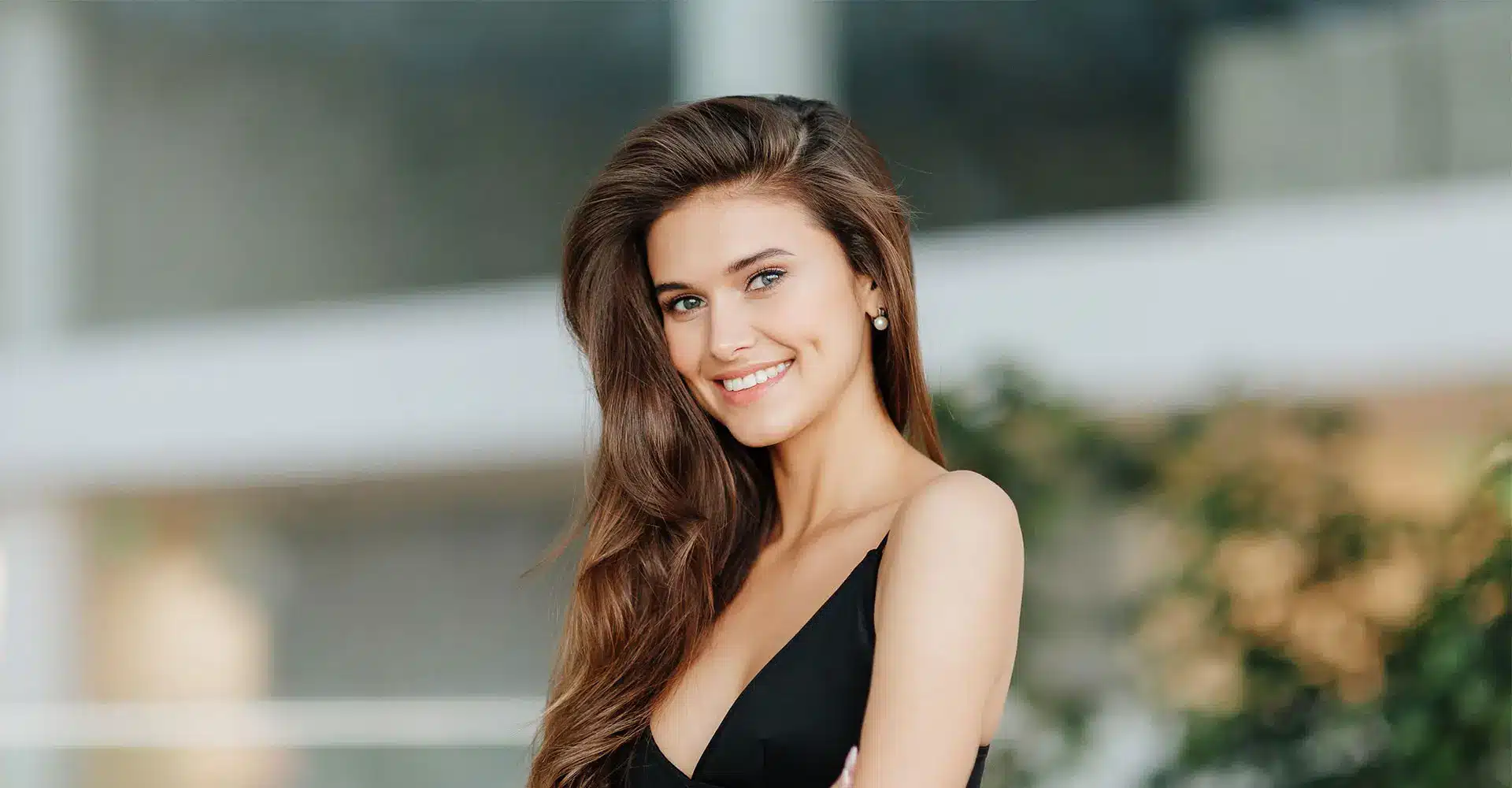 a woman with long brown hair and a bright smile is standing outdoors she is wearing a sleeveless black top and has her arms crossed looking radiant after her recent brow lift the background is blurred with greenery and a building visible