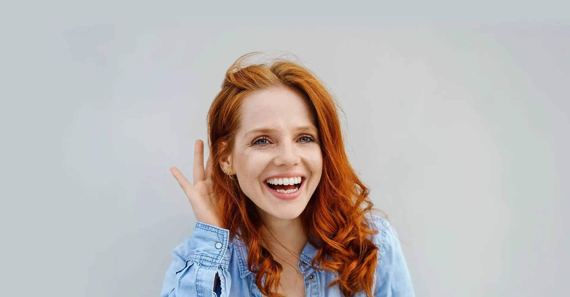 a woman with long curly red hair and wearing a light blue shirt smiles brightly while holding one hand next to her ear possibly highlighting the results of an earlobe reduction she stands against a plain light colored background
