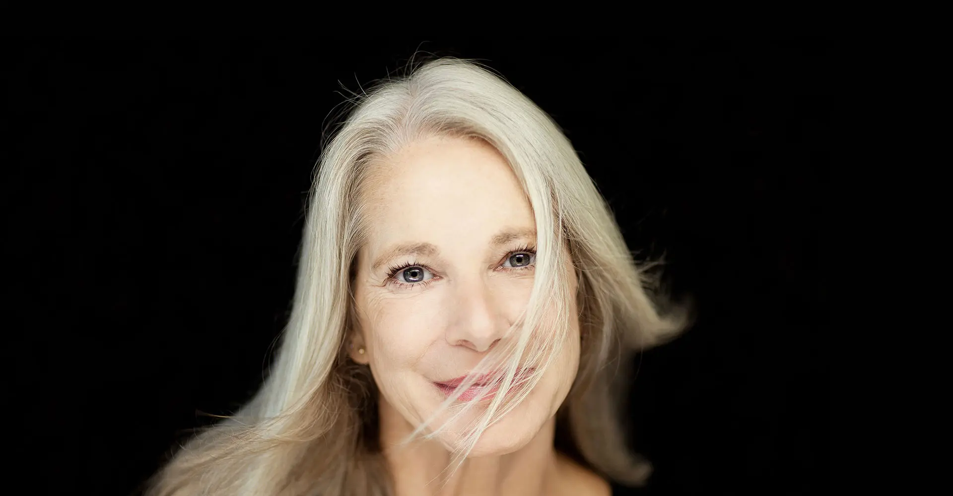 a woman with long gray hair possibly enhanced by facial surgery smiles softly at the camera her hair gently flows across her face partially covering her left eye she is set against a dark black background which contrasts with her light hair and bright complexion