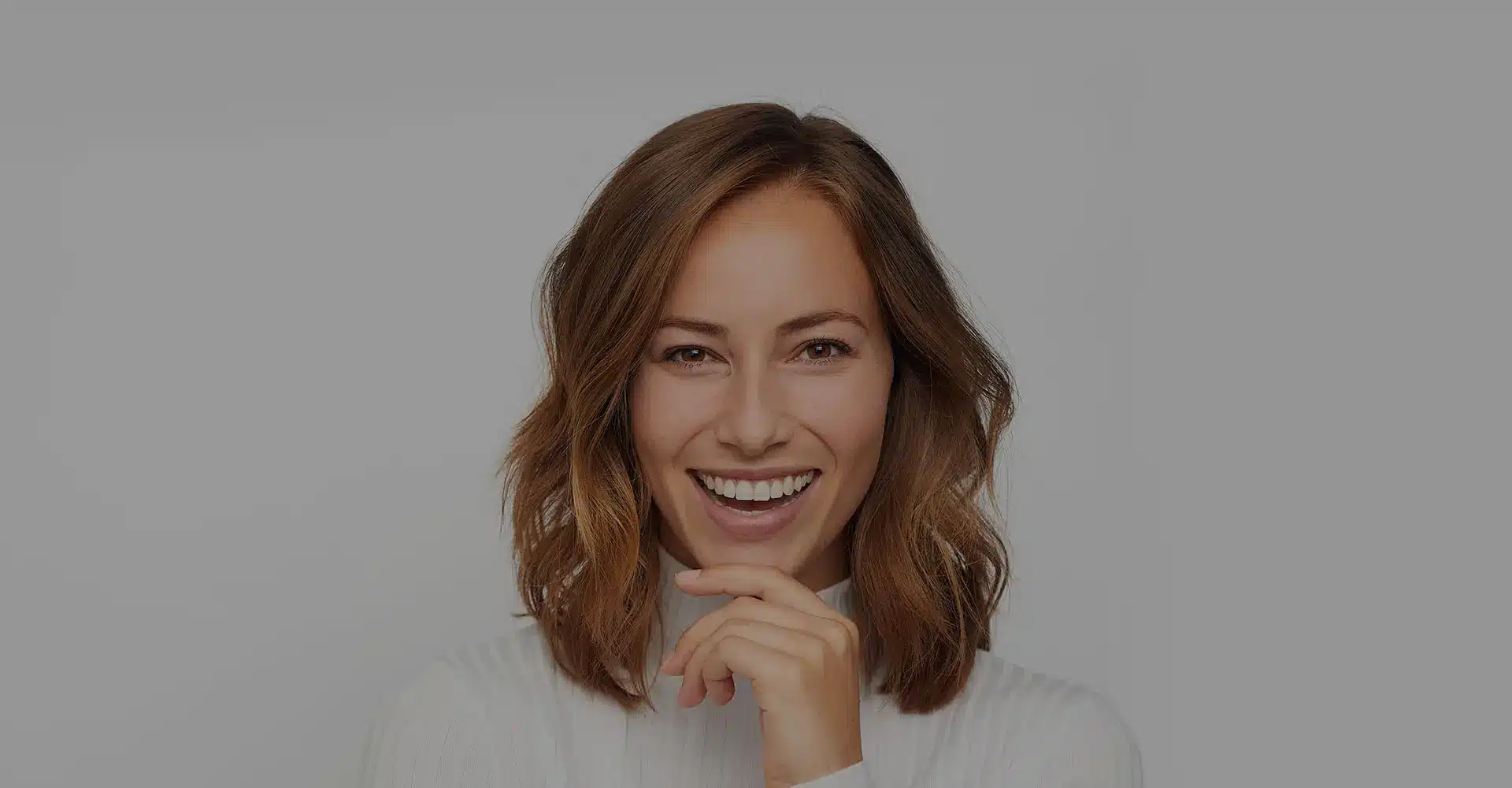 a woman with shoulder length brown hair smiles brightly while touching her chin with her right hand she is wearing a white long sleeved top and standing against a plain light gray background myellevate offers her the perfect opportunity to shine brighter in any setting