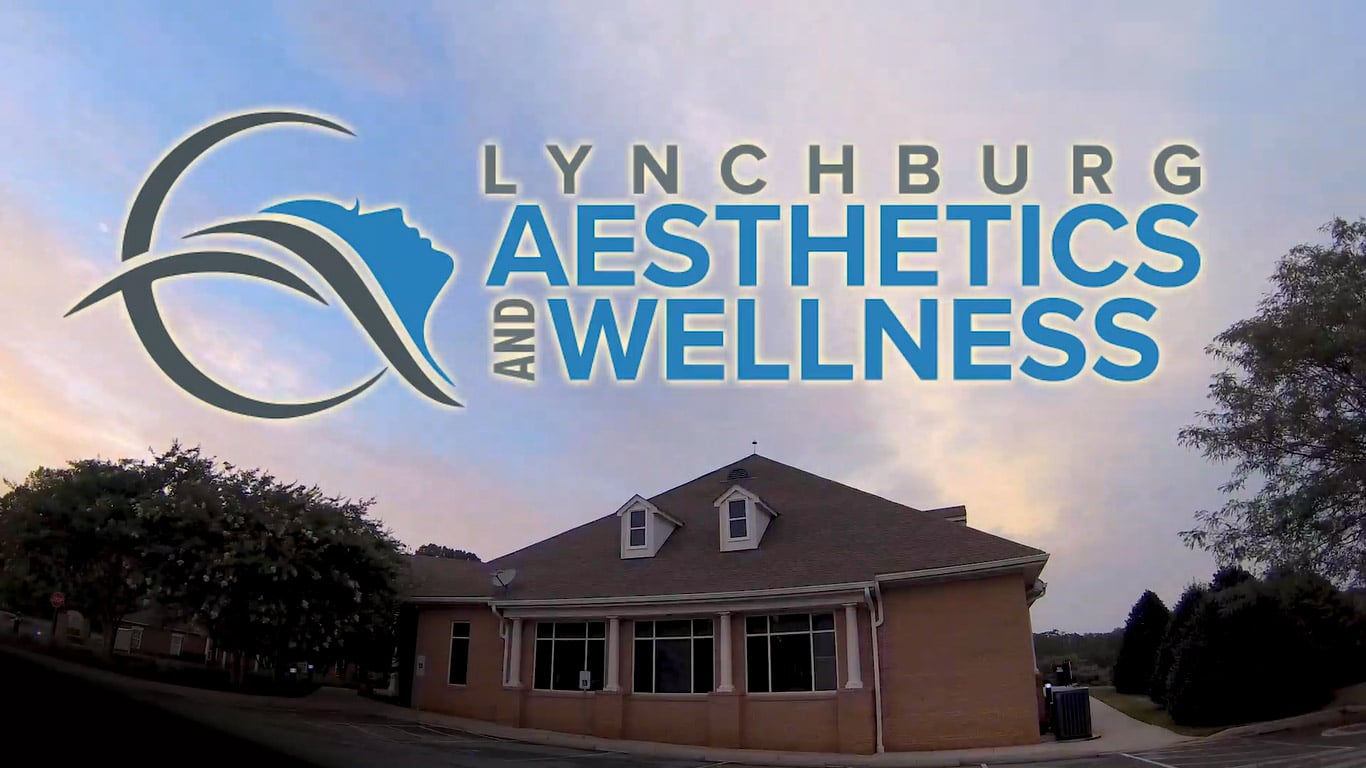 a brick building with a gabled roof sits under a sky with pastel clouds large blue and white text overlaid on the sky reads lynchburg aesthetics and wellness with an abstract logo featuring a curved line and a profile of a face led by dr william carvajal trees are visible on the sides