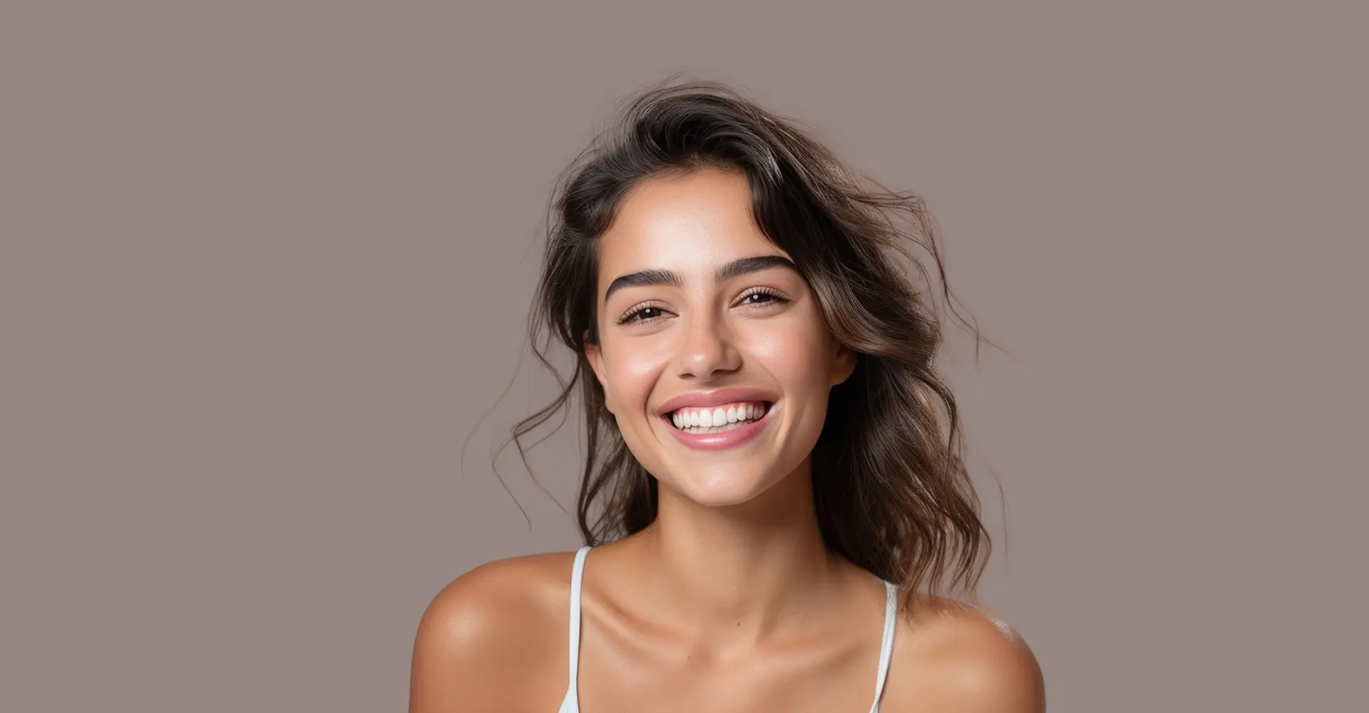 a young woman with wavy brown hair smiles brightly at the camera radiating happiness and confidence she is wearing a white tank top against a solid neutral background her recent bbl has enhanced her natural beauty adding to her radiant glow