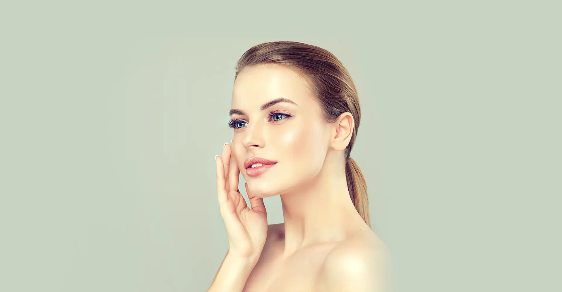a woman with fair skin and blonde hair pulled back into a ponytail is gazing to the side with her hand gently touching her cheek she appears serene showcasing natural beauty with minimal makeup and perfectly shaped eyebrows set against a neutral gray background