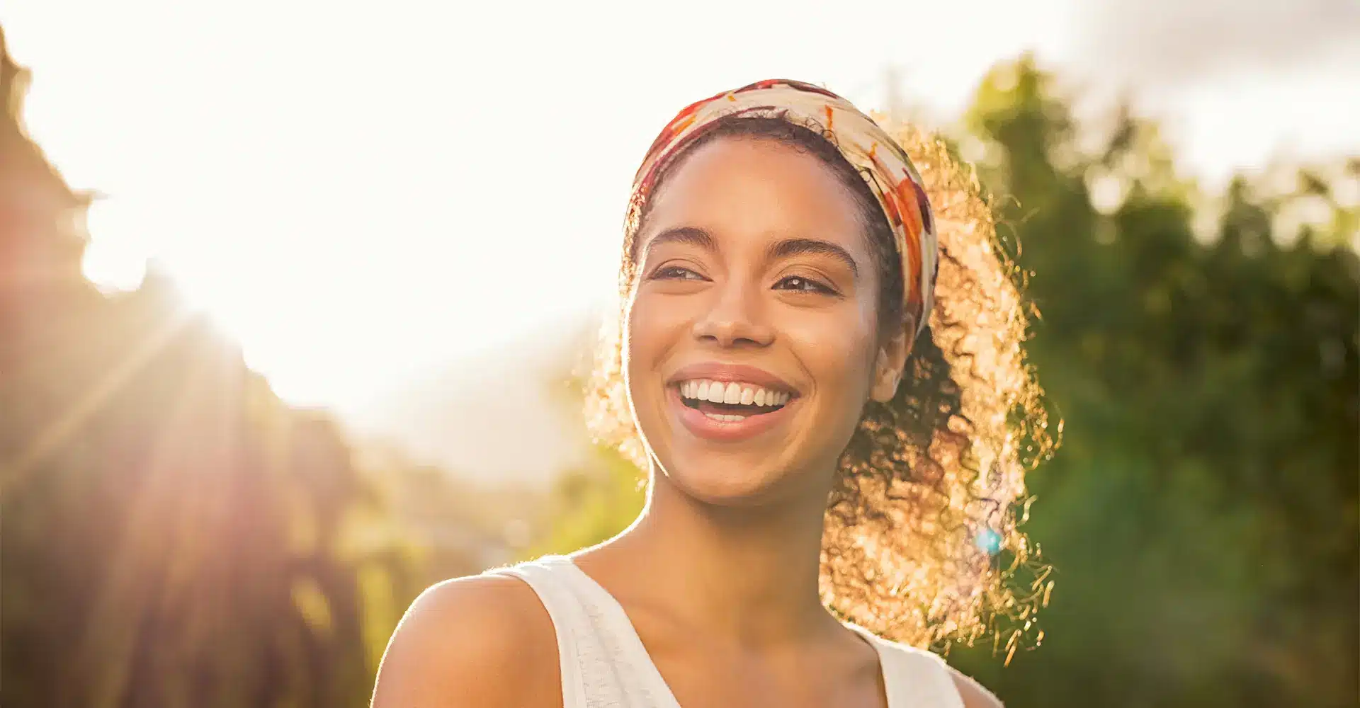 a young woman with curly hair wearing a headband smiles brightly while standing outdoors against a backdrop of greenery and warm sunlight she is dressed in a light colored top exuding natural beauty that complements her radiant skin possibly enhanced by chemical peels