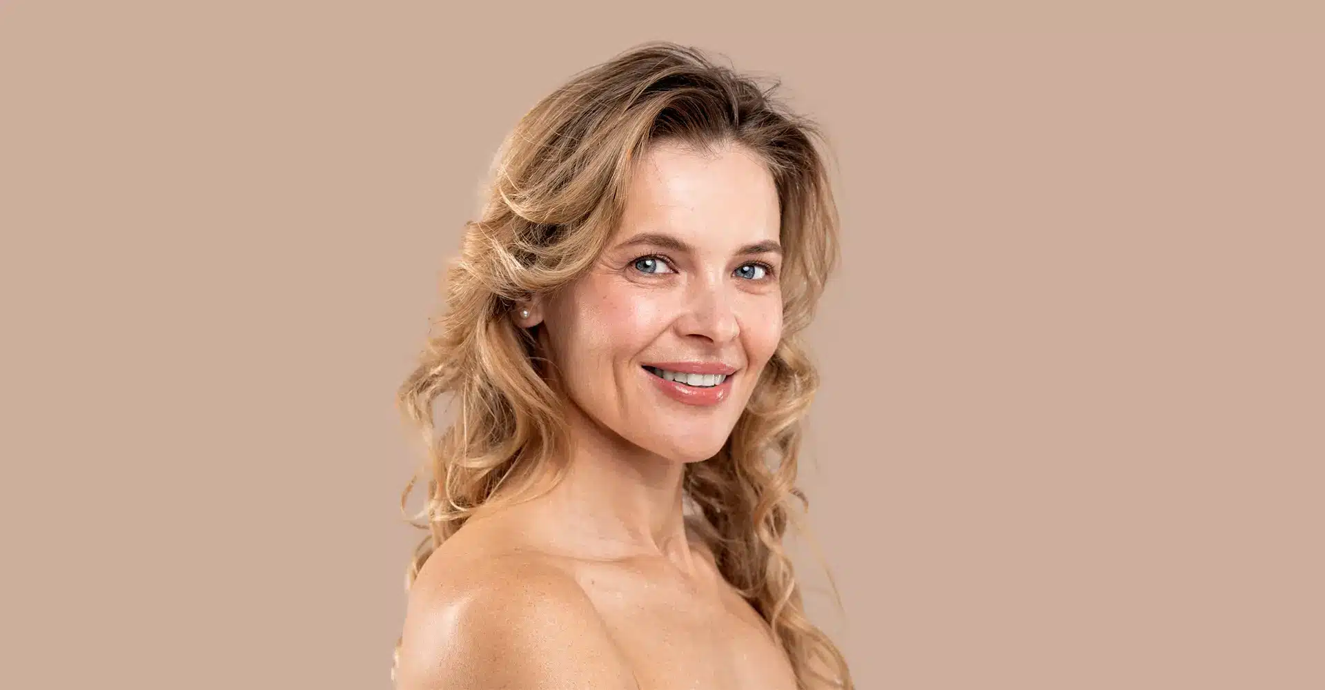 a woman with long curly blonde hair smiling at the camera in addition to her radiant beauty she recently underwent co laser resurfacing enhancing her glow she is posed against a plain beige background and her shoulders are bare