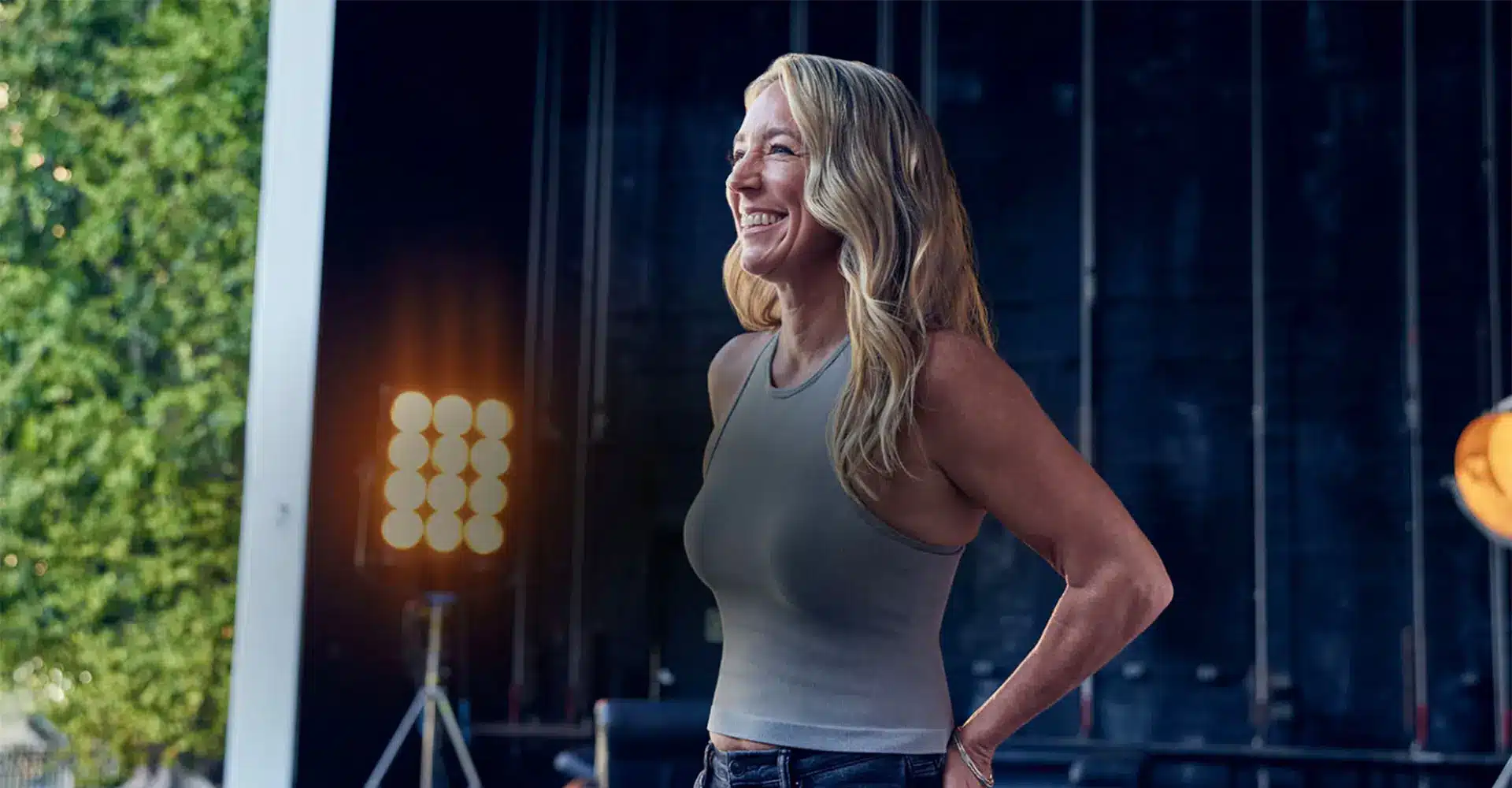 a woman with long blond hair stands outdoors near a large tree and stage lighting equipment she smiles and wears a gray sleeveless top and jeans showcasing the results of her recent coolsculpting session the background includes a dark wall with what appears to be stage rigging equipment