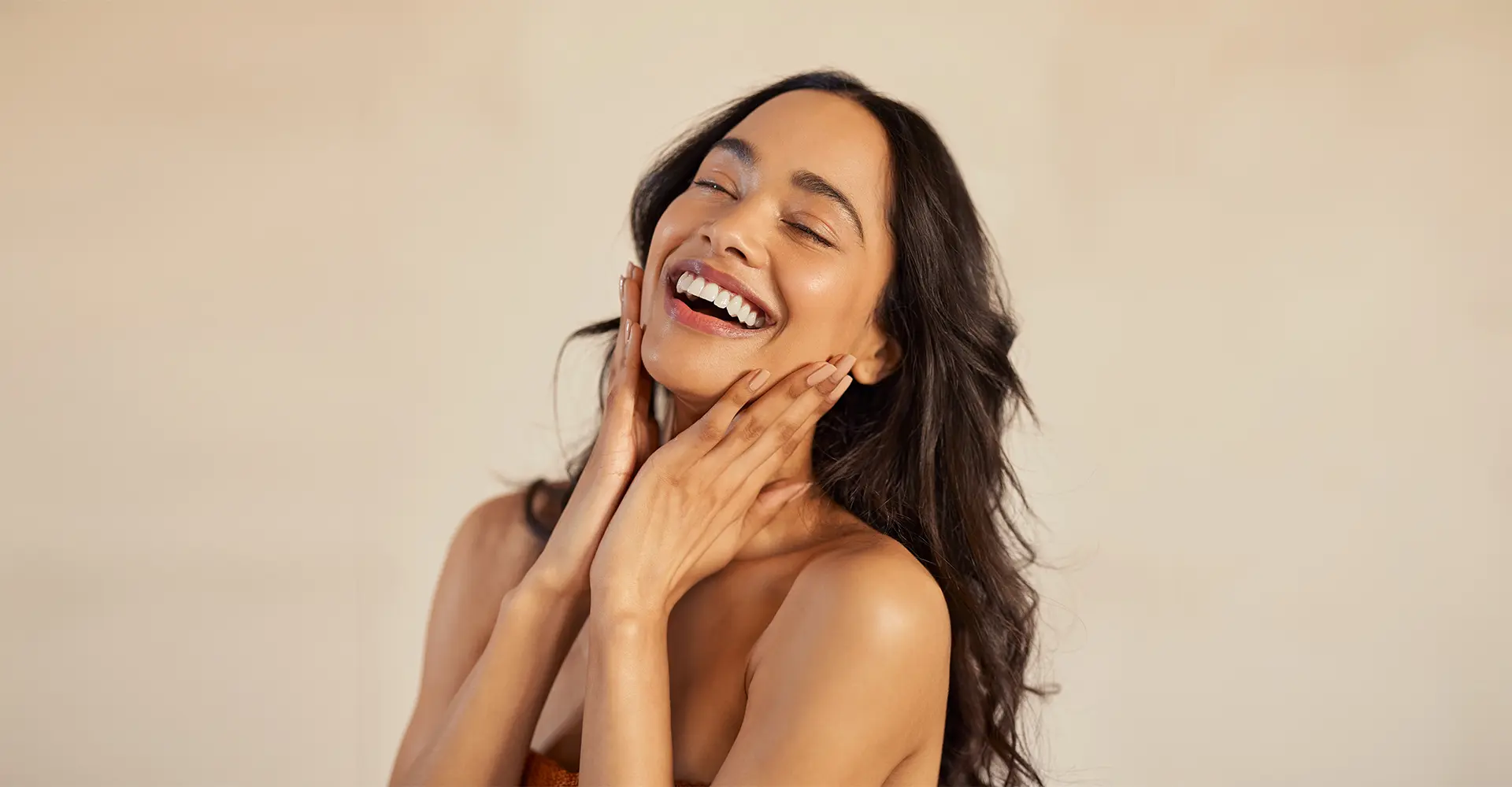 a woman with long dark hair and a radiant smile tilts her head back slightly touching her face with both hands eyes closed she appears to be laughing the plain softly lit background enhances the serene feel of the image embodying the joy of a refreshing skincare treatment