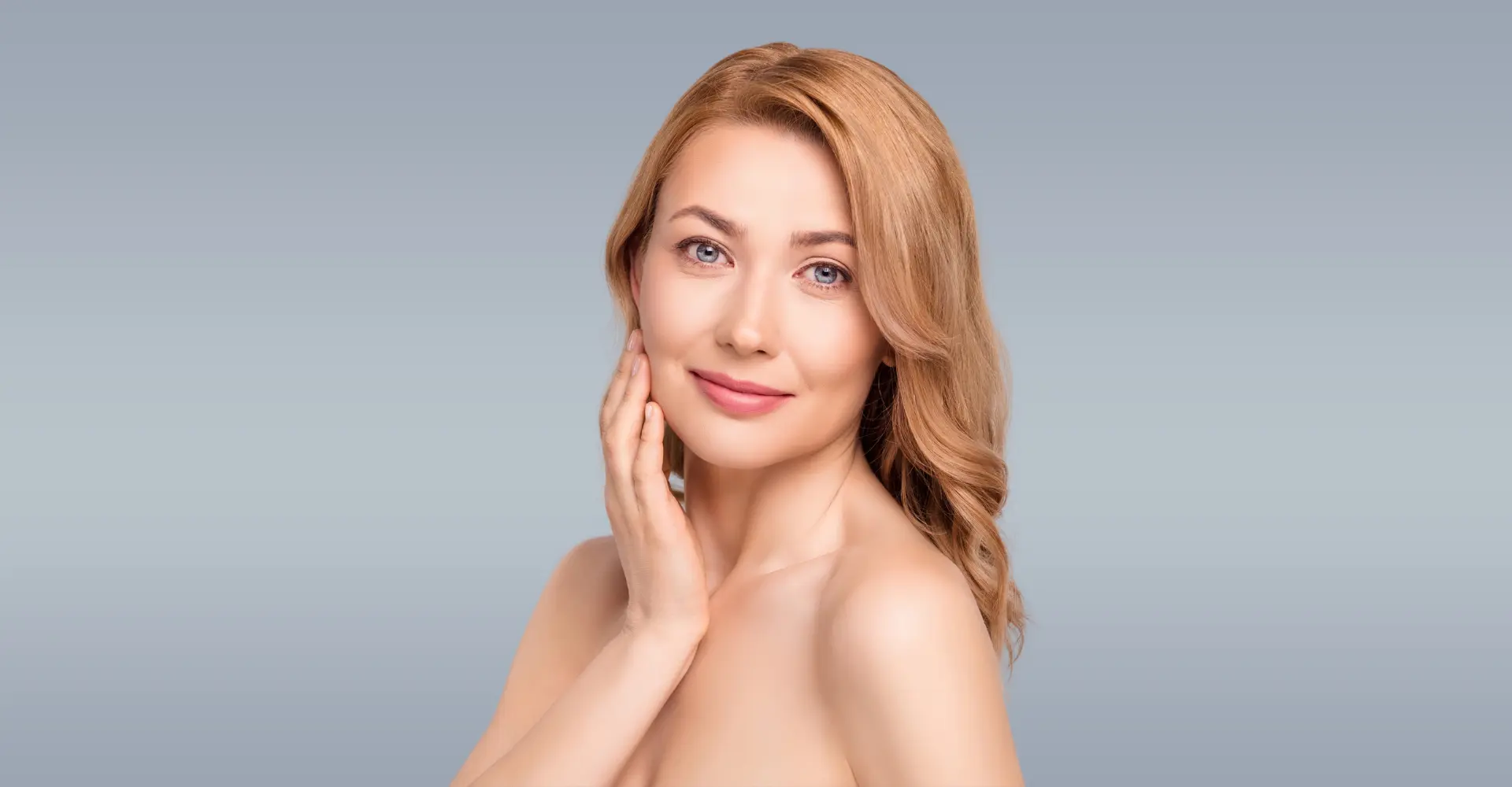 a woman with fair skin and long wavy blonde hair is smiling softly she has her right hand gently touching her cheek showing off radiant skin that looks like it has benefited from an erbium yag laser treatment the background is a plain light gradient shifting from light blue to gray she has minimal makeup and a serene expression