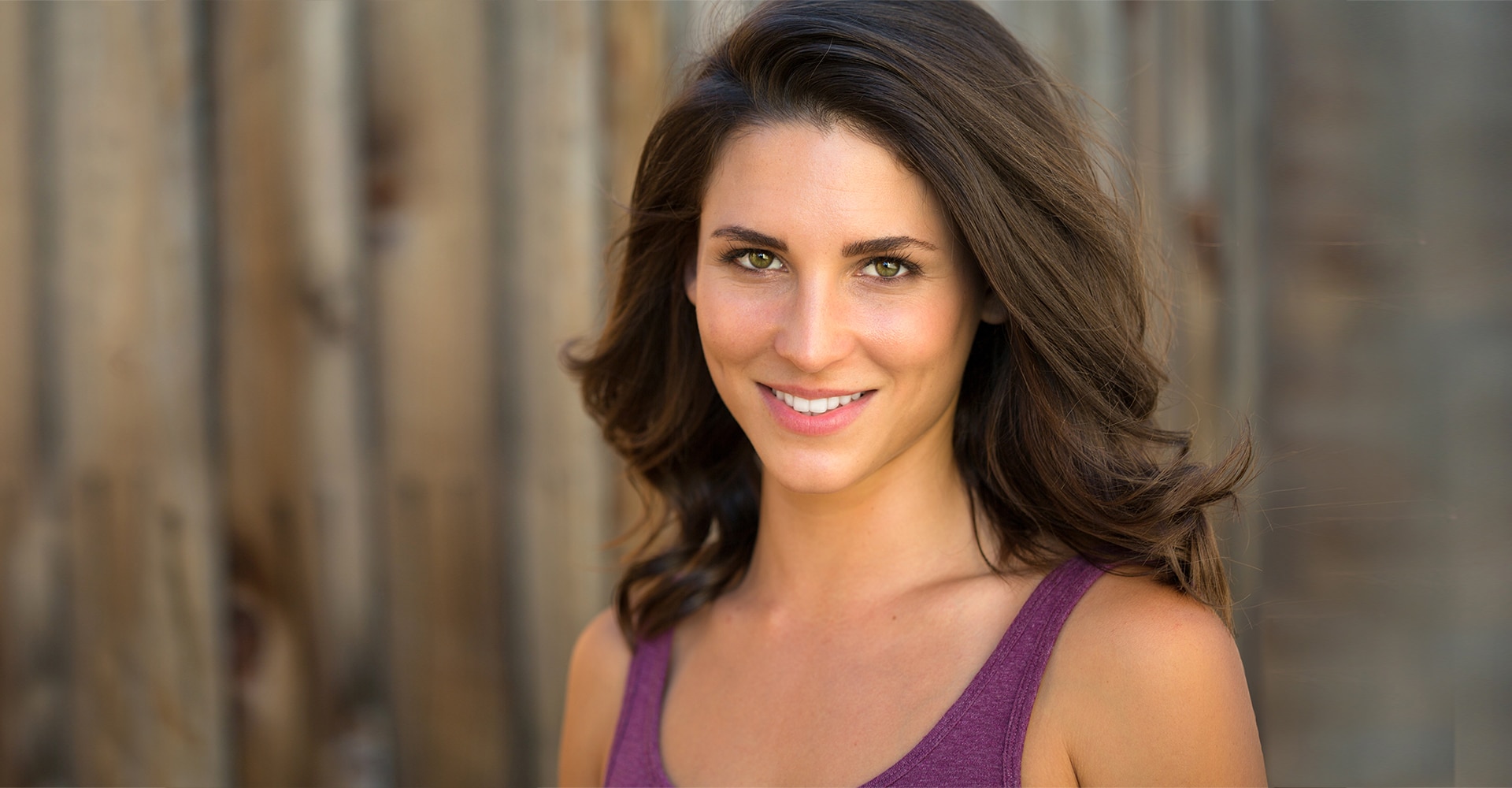 a woman with long brown hair and green eyes is smiling wearing a purple sleeveless top the background consists of wooden planks creating a serene setting that highlights her glowing skin enhanced by her recent hydrafacial treatment