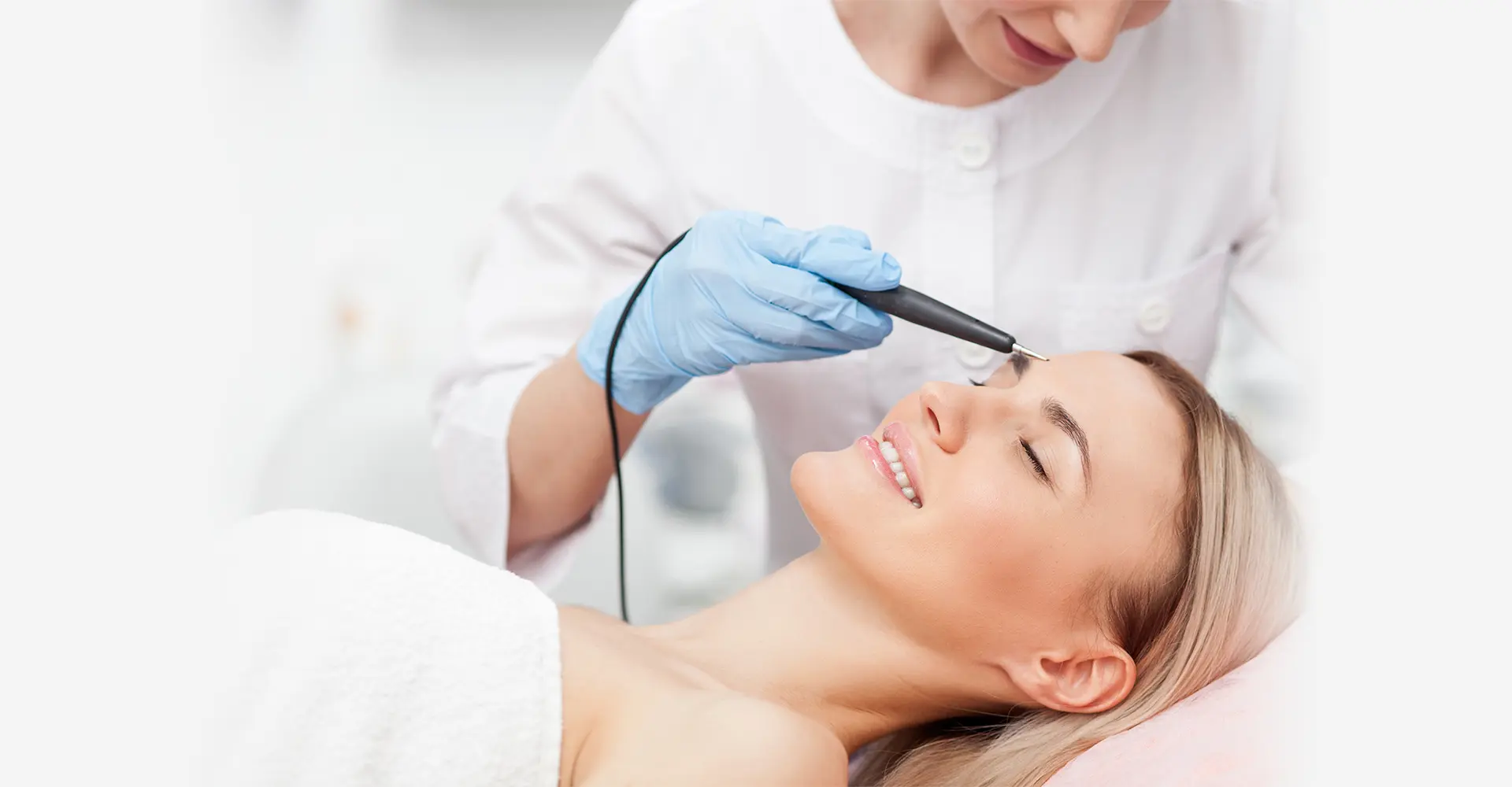 a woman lies on a treatment table with her eyes closed smiling while a healthcare professional in a white uniform and blue gloves holds a device to her forehead the setting appears to be a medical or spa facility possibly providing laser hair removal services