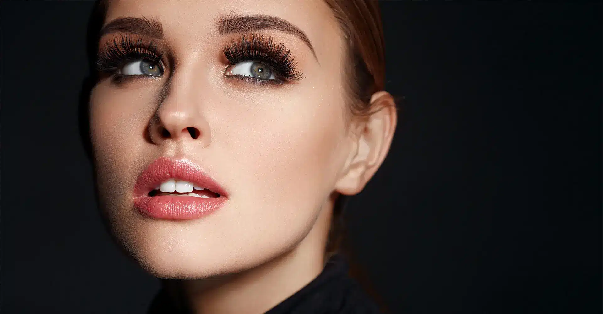 a close up of a person with fair skin and light pink lips looking upward they have long voluminous eyelashes thanks to an eyelash enhancement their perfectly arched eyebrows complete the look the background is a dark solid color creating a dramatic contrast with their well lit face