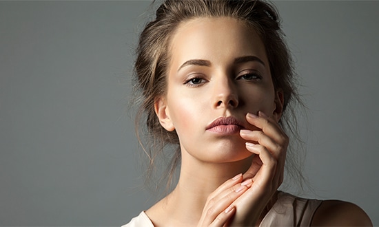 a woman with light skin and brown hair loosely tied back poses against a gray background gazing at the camera with a neutral expression her right hand gently touches her chin and her left hand rests on her collarbone embodying the elegance often seen in beauty treatments and skincare promotions