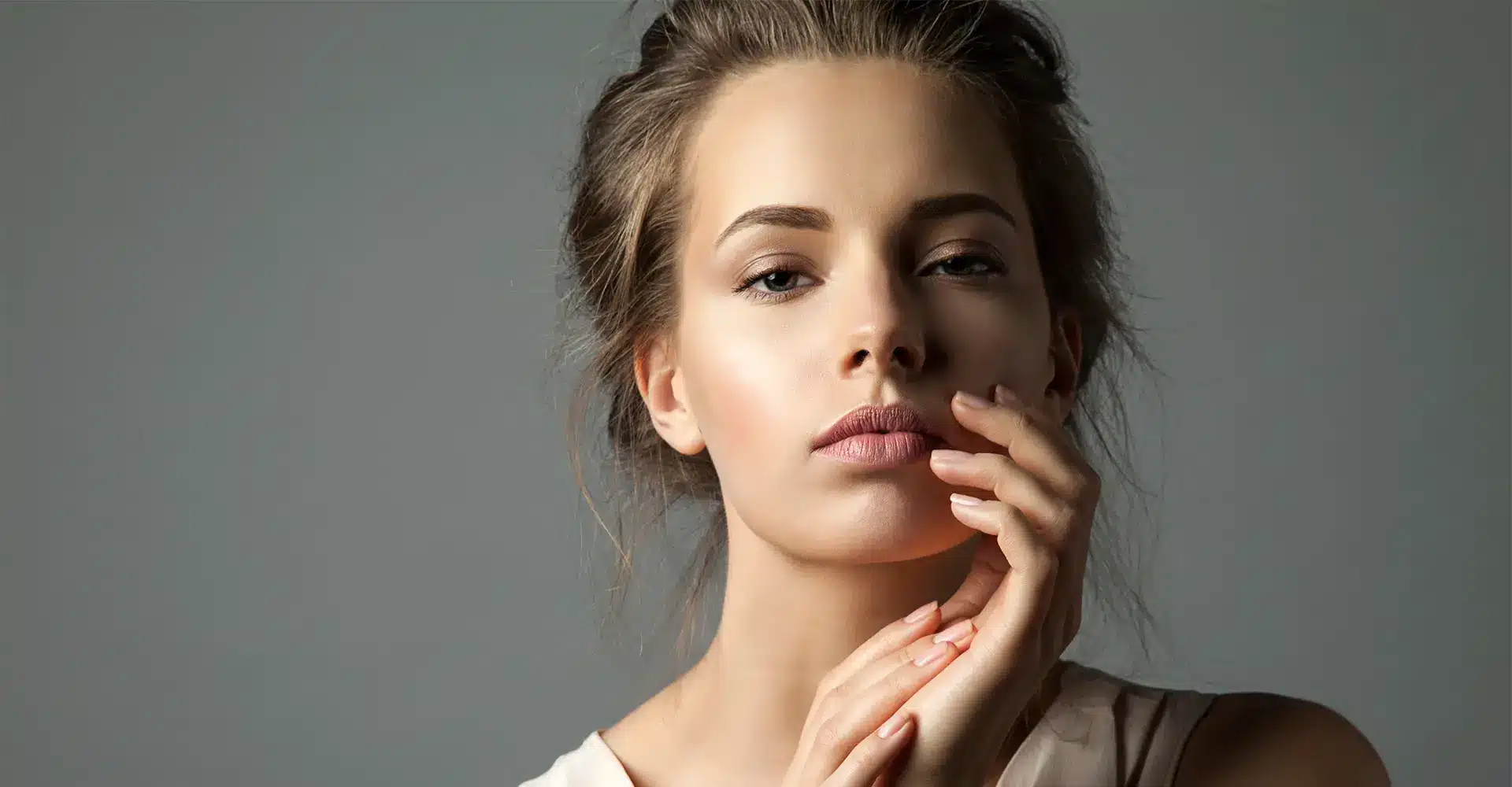 a young woman with light skin and brown hair pulled back gazes directly at the camera with a neutral expression she has her hand gently touching the side of her face showcasing her beautifully enhanced brows from a professional brow tinting session she is wearing a sleeveless light colored top against a plain gray background