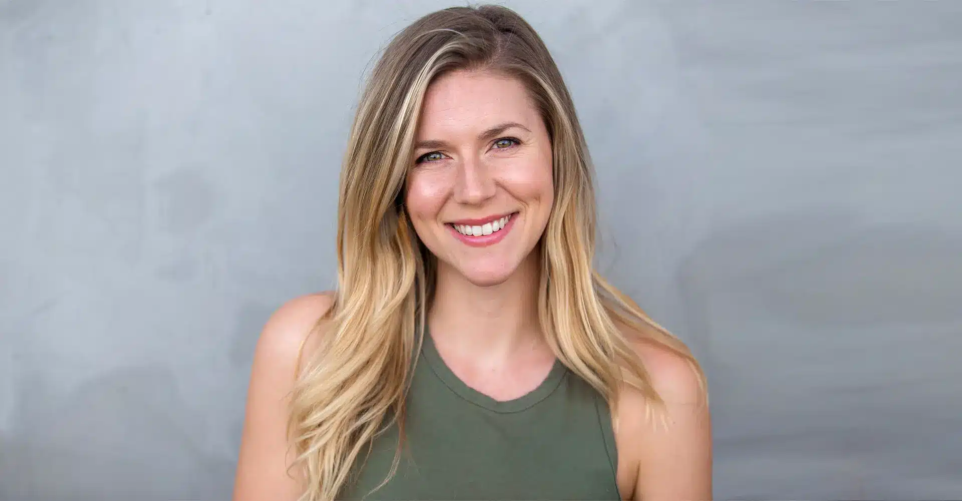 a woman with long blonde hair wearing a sleeveless green top smiles at the camera against a plain light gray background showcasing the results of her recent hair restoration
