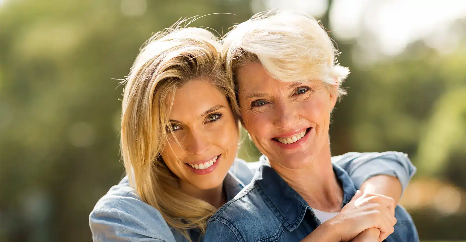 two smiling women with blonde hair dressed in casual blue tops pose closely outdoors the younger woman has her arm around the older woman showing a warm affectionate bond their radiant skin glows in the sunlight and the background features greenery creating a cheerful setting