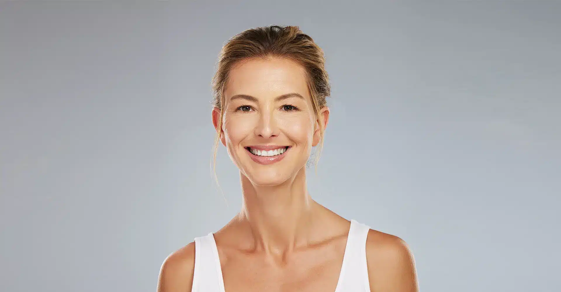 a woman with pulled back hair smiles at the camera she is wearing a white sleeveless top exuding confidence as she stands against a neutral grey background highlighting the transformative effects of morpheus