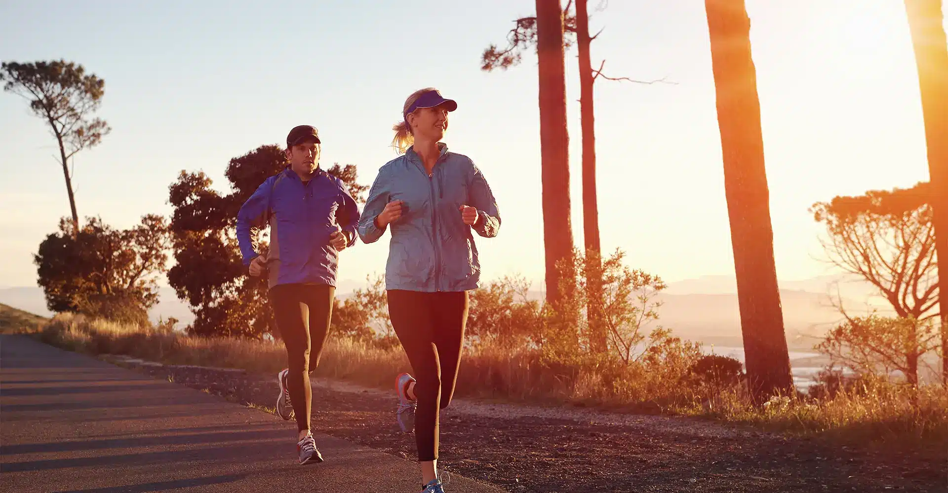 two individuals are jogging on an outdoor trail at sunrise embracing the essence of wellness dressed in athletic wear including caps and jackets they move along the path lined with tall trees the sun casts a warm glow creating long shadows and illuminating the morning landscape