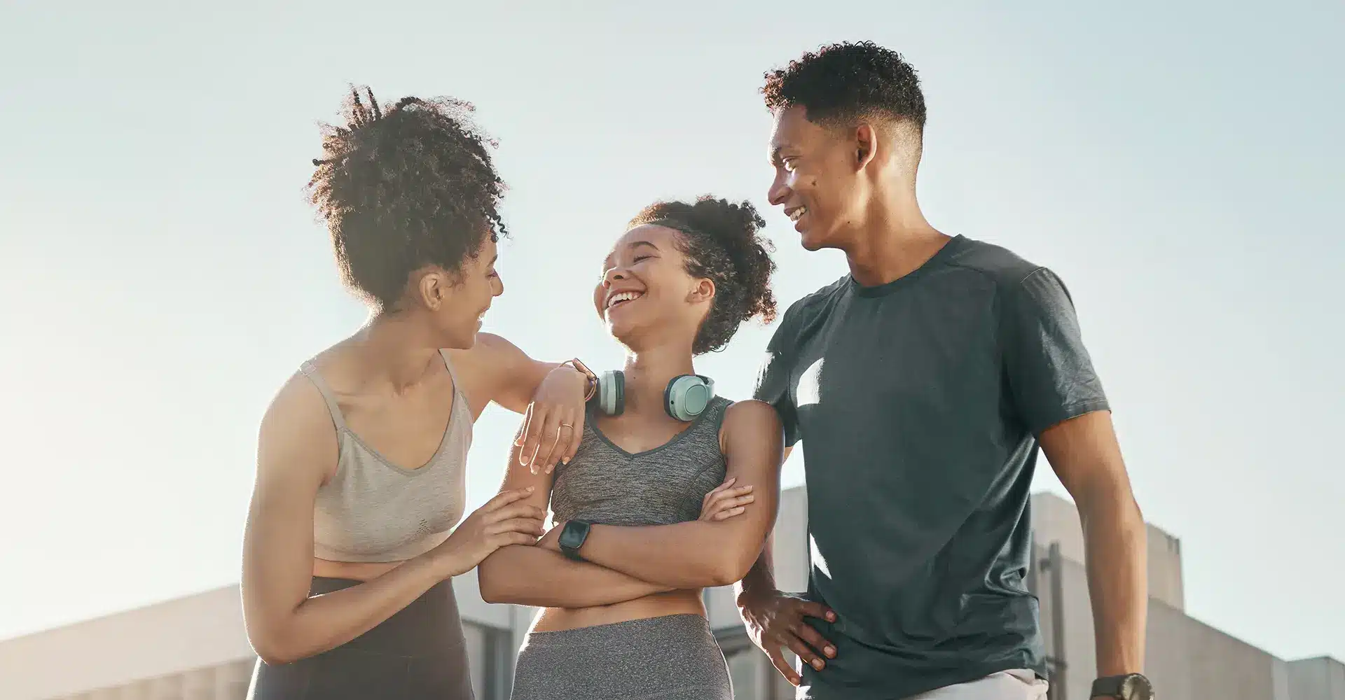 three people enjoying each others company outdoors on a sunny day two women dressed in athletic wear stand on either side of a man the woman on the left has her arm around the shoulders of the woman in the middle who is smiling and wearing headphones around her neck after an emsculpt session