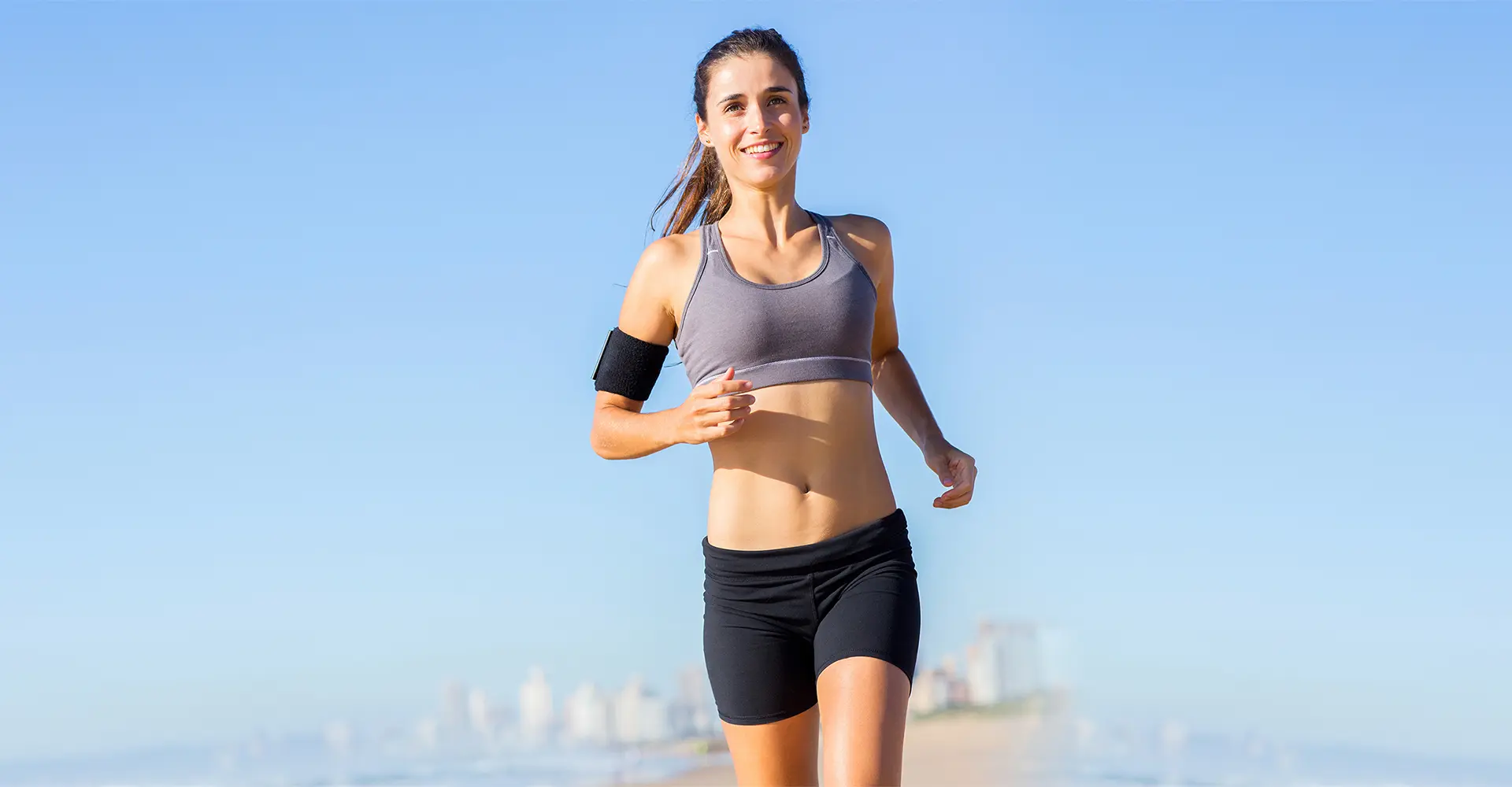 a woman in athletic attire is jogging by the beach showcasing how fitness routines can evolve she is wearing a sports bra shorts and an armband with her hair pulled back the background features a clear blue sky and distant cityscape she looks happy and energized while running