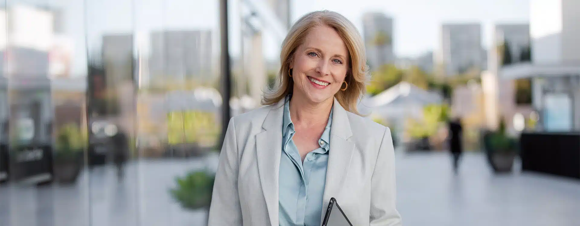 a woman with blonde hair wearing a light grey blazer and a light blue shirt stands outdoors smiling she holds a tablet in her hand the background features modern buildings a glass wall and greenery